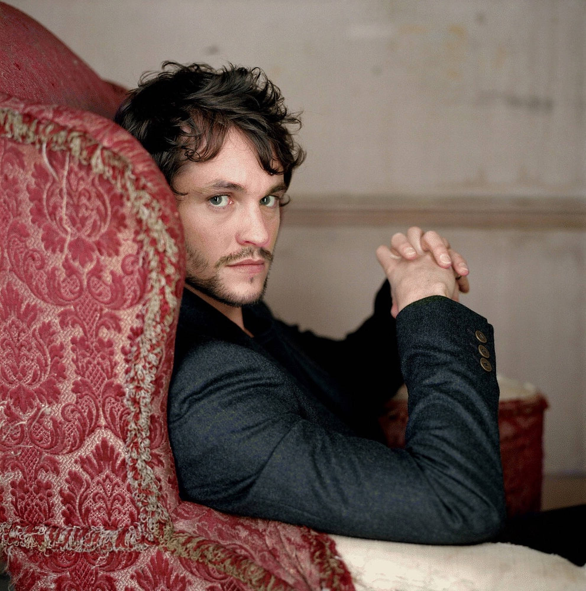 Hugh Dancy 桌布 壁紙 — 一名黑髮綠眼的男子在柔和光線的房間裡靠在一張紅色錦緞椅子上。