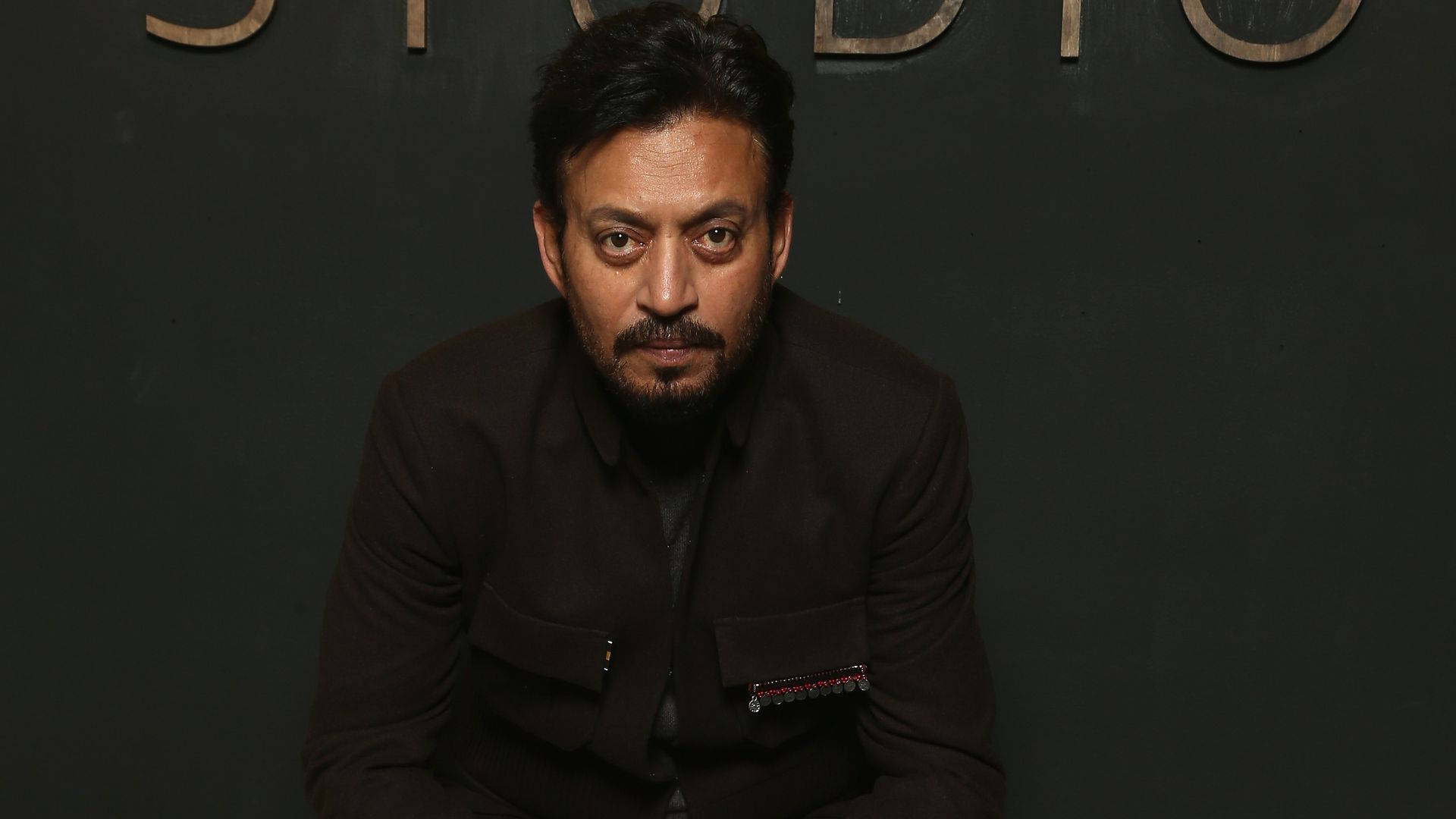 Irrfan Khan 桌布 壁紙 — 一名黑髮鬍鬚男穿著深棕色外套，背景是黑色牆壁上有金色字體。