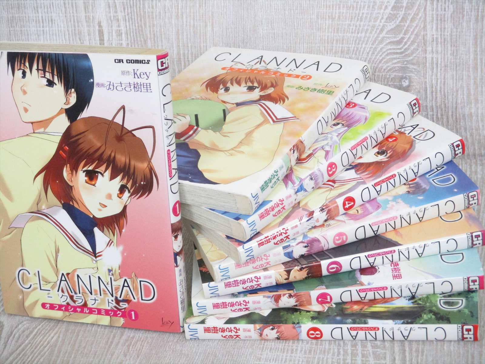 Clannad 桌布 壁紙 — 一堆Clannad漫畫卷，封面採用動漫風格，配有校服和柔和粉彩背景。