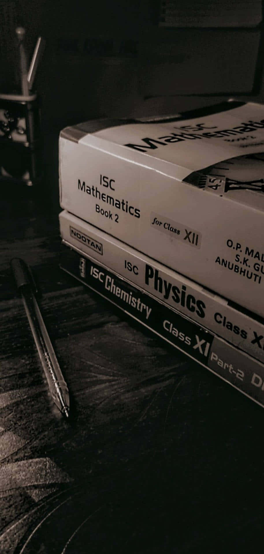 Dark Book Aesthetic 桌布 壁紙 — 一堆ISC物理和數學教科書（適用於XI和XII年級），旁邊放著一支筆，放在深色木桌上。
