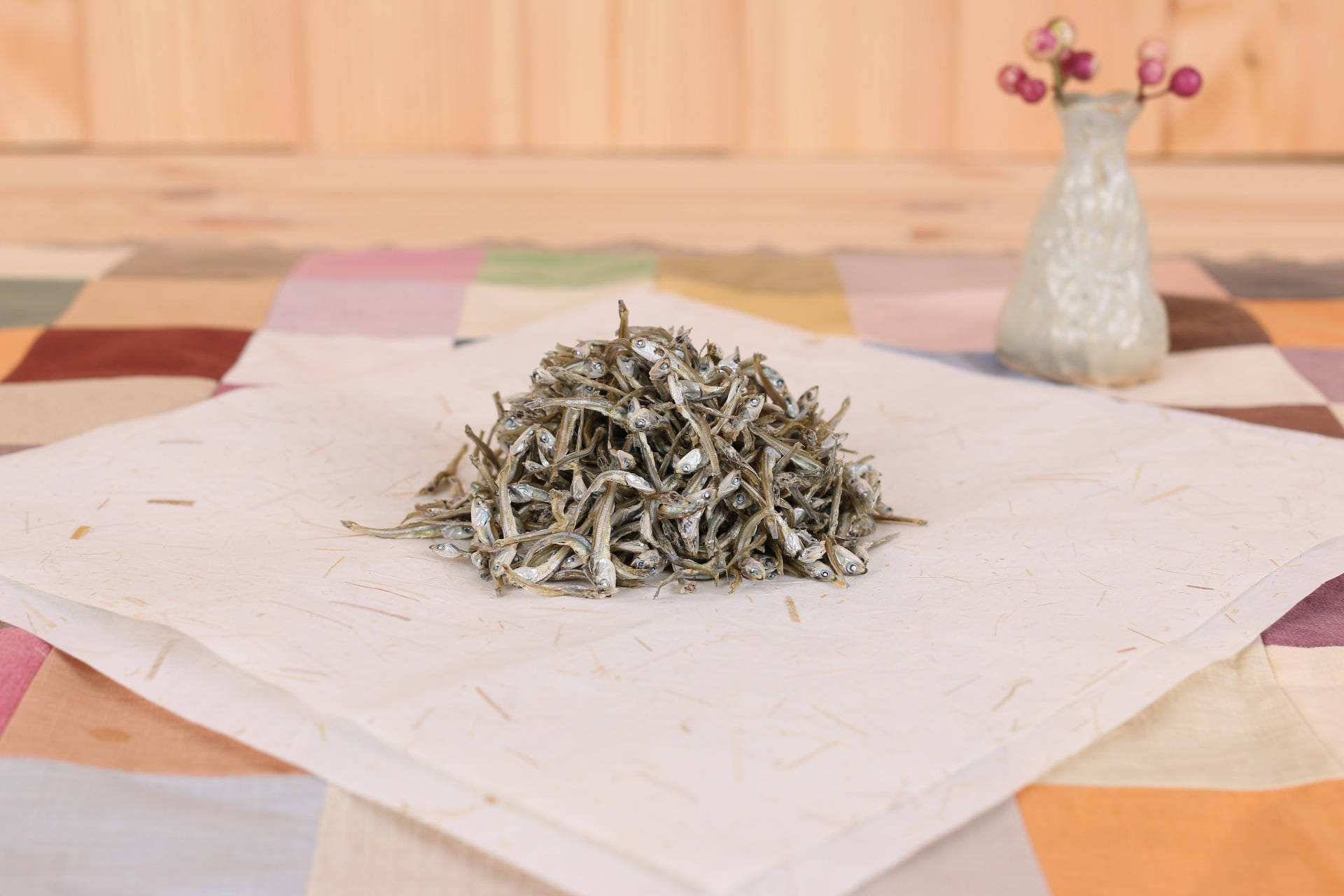 魚 桌布 壁紙 — 一堆乾銀魚放在有紋理的紙上，紙下是色彩繽紛的拼布桌布，背景有一個陶瓷花瓶插著粉紅色的漿果。