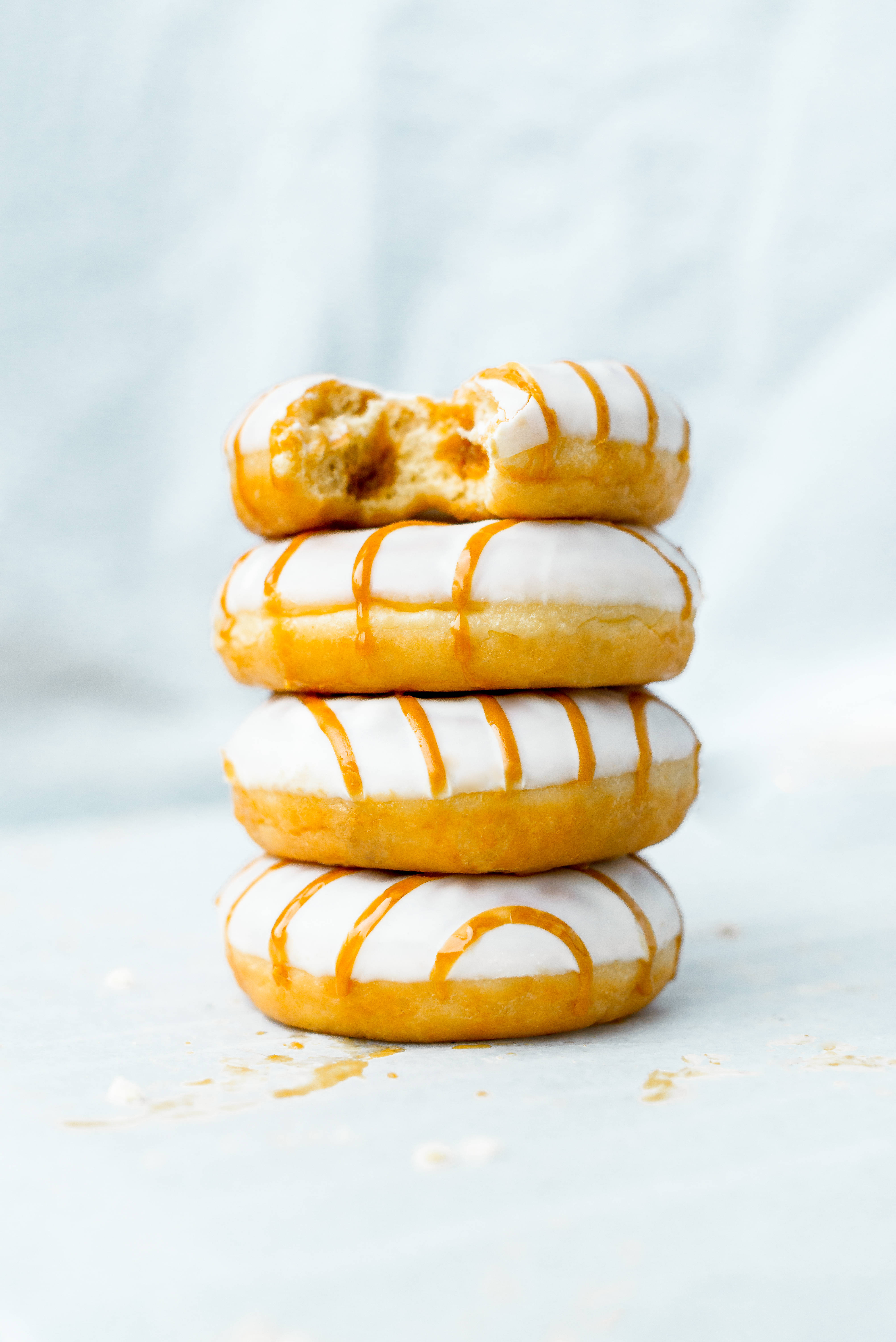 Pastry 桌布 壁紙 — 一堆四個糖霜甜甜圈，淋上焦糖，其中一個被咬了一口，露出柔軟的麵包屑，背景是柔和的藍色。