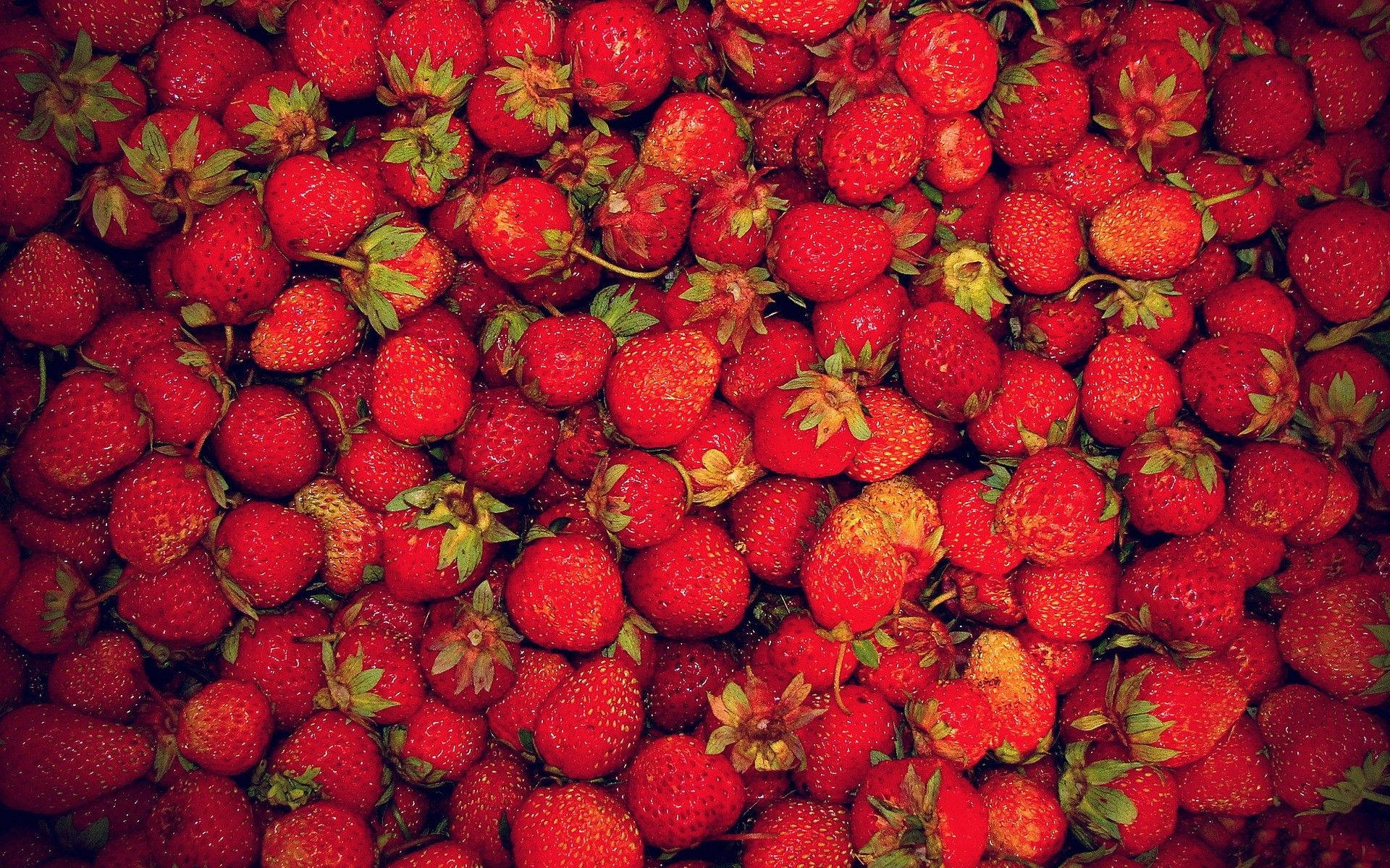 Strawberry Aesthetic 桌布 壁紙 — 一堆密集的成熟紅草莓，帶有綠色頂部和細小種子，在溫暖燈光下閃閃發光。