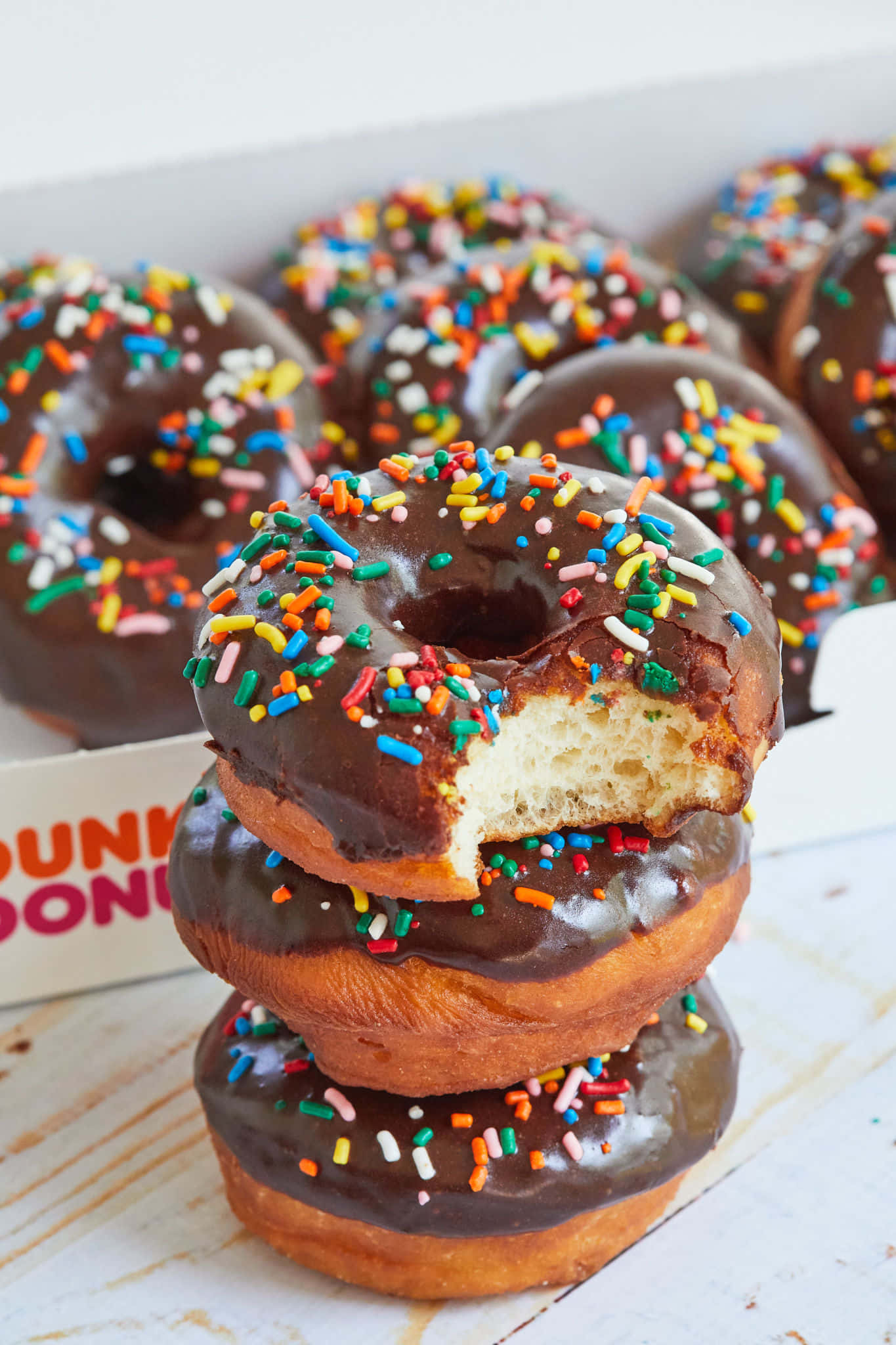 Dunkin Donuts 桌布 壁紙 — 一堆巧克力糖霜甜甜圈，上面有彩色糖粒，其中一個被咬了一口，露出柔軟蓬鬆的內部。