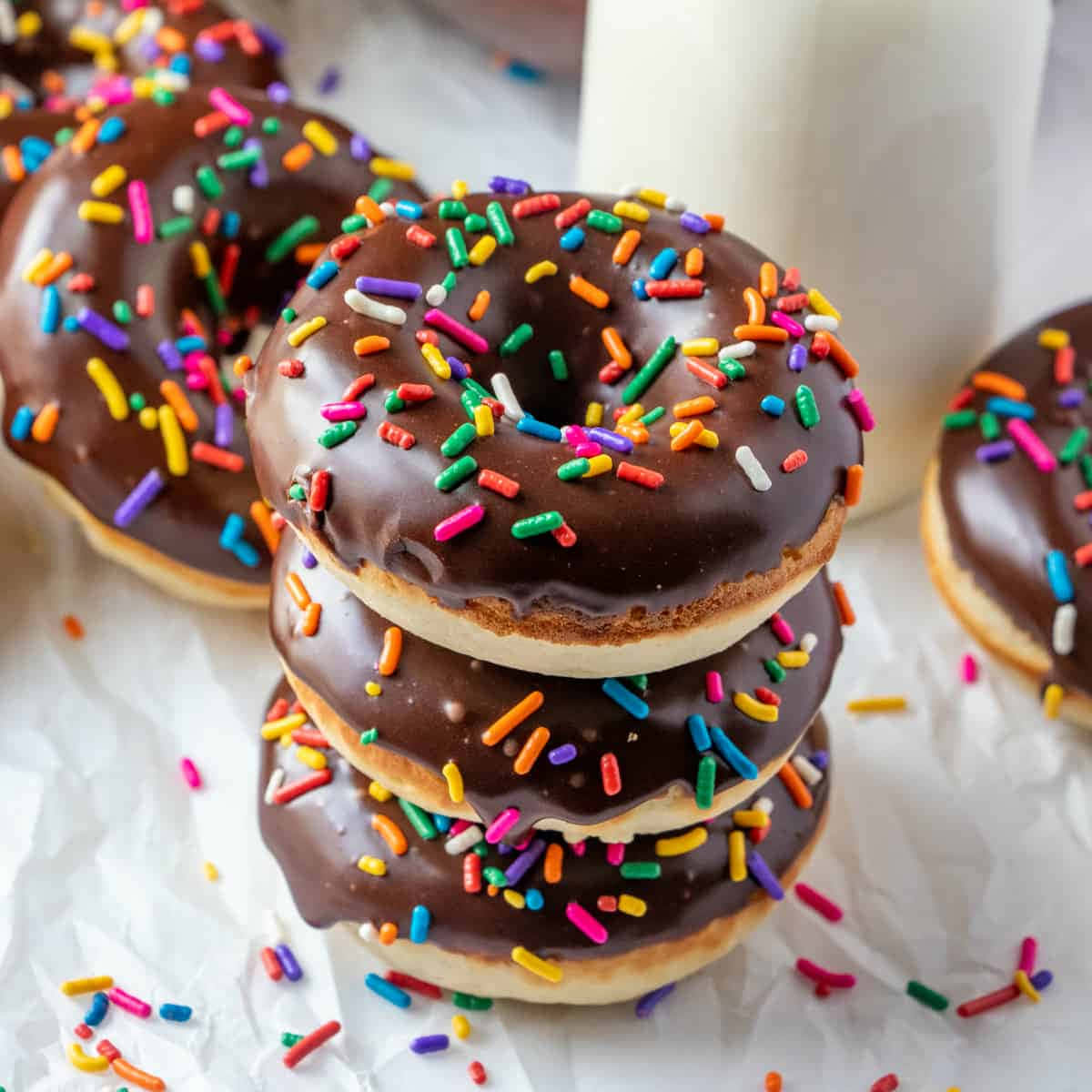 Glazed Donut 桌布 壁紙 — 一堆巧克力釉面甜甜圈，上面撒滿彩虹糖針，放在皺巴巴的烘焙紙上，旁邊有一杯牛奶。