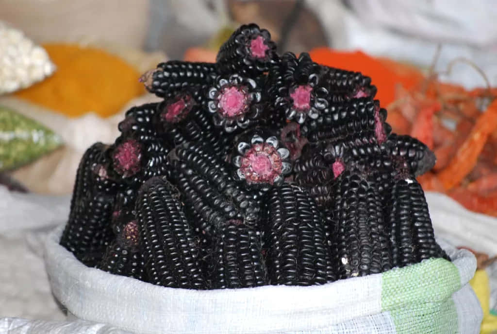Purple Corn 桌布 壁紙 — 一堆帶有粉紅色花心的光澤黑玉米棒放在鋪有白布的籃子裡。