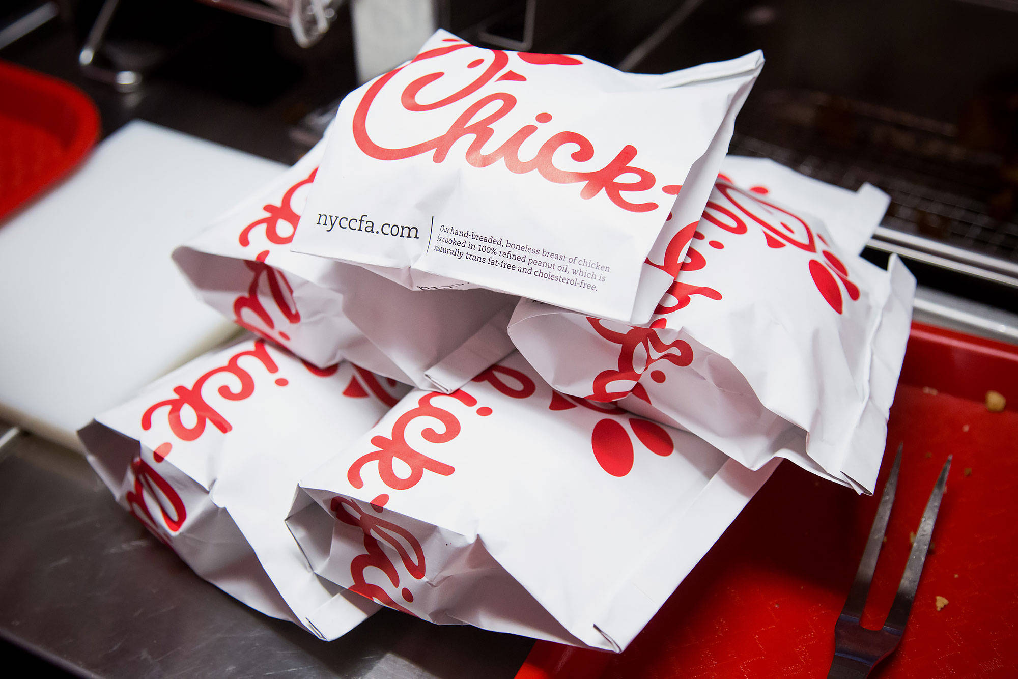 chick 桌布 壁紙 — 一堆帶有紅色標誌的白色Chick-fil-A紙袋放在快餐廚房的金屬叉旁邊的紅色托盤上。