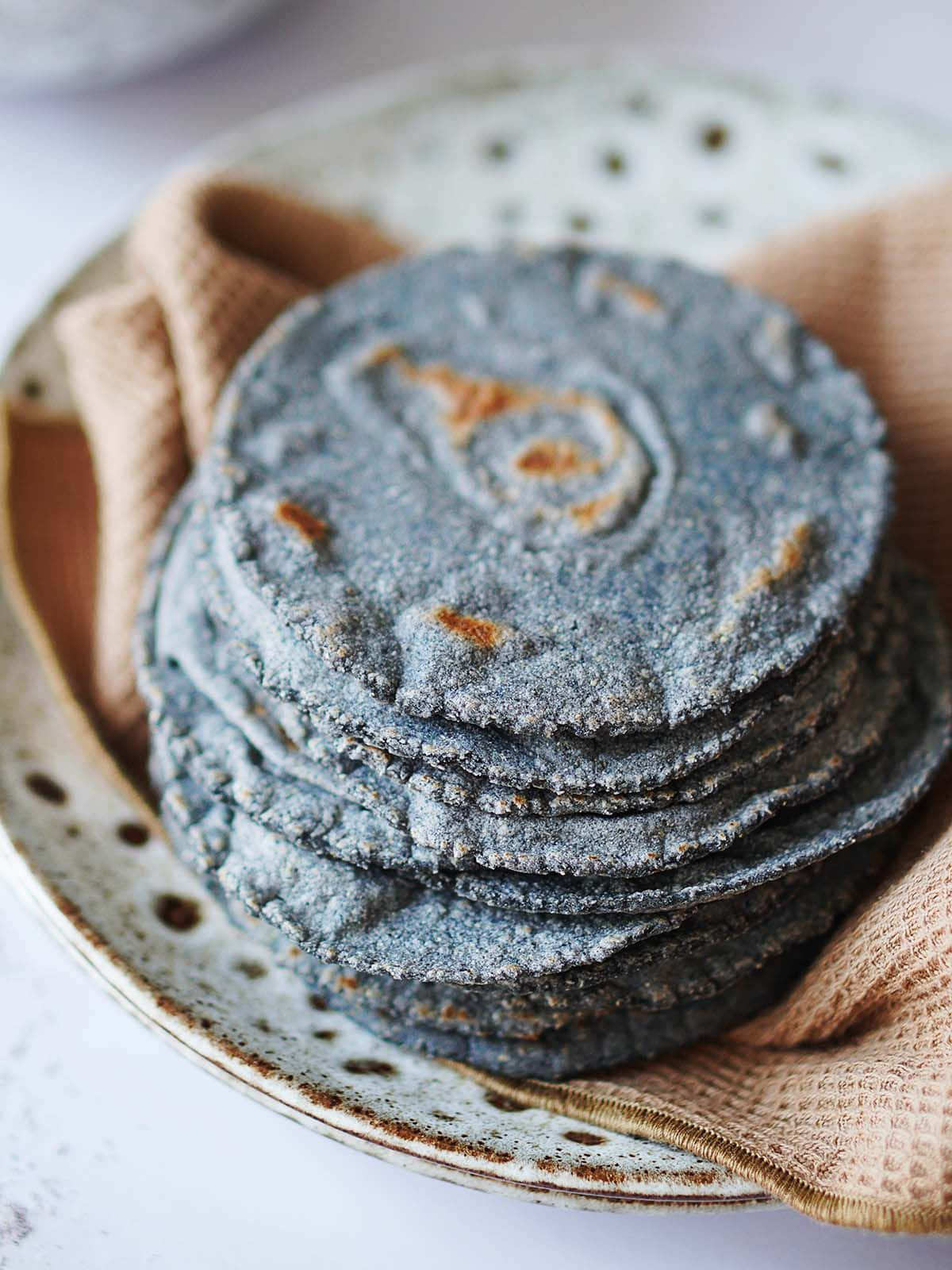Blue Chips 桌布 壁紙 — 一堆帶有金色烤痕的鄉村藍玉米餅，放在鋪有米色布的斑點陶瓷盤上。