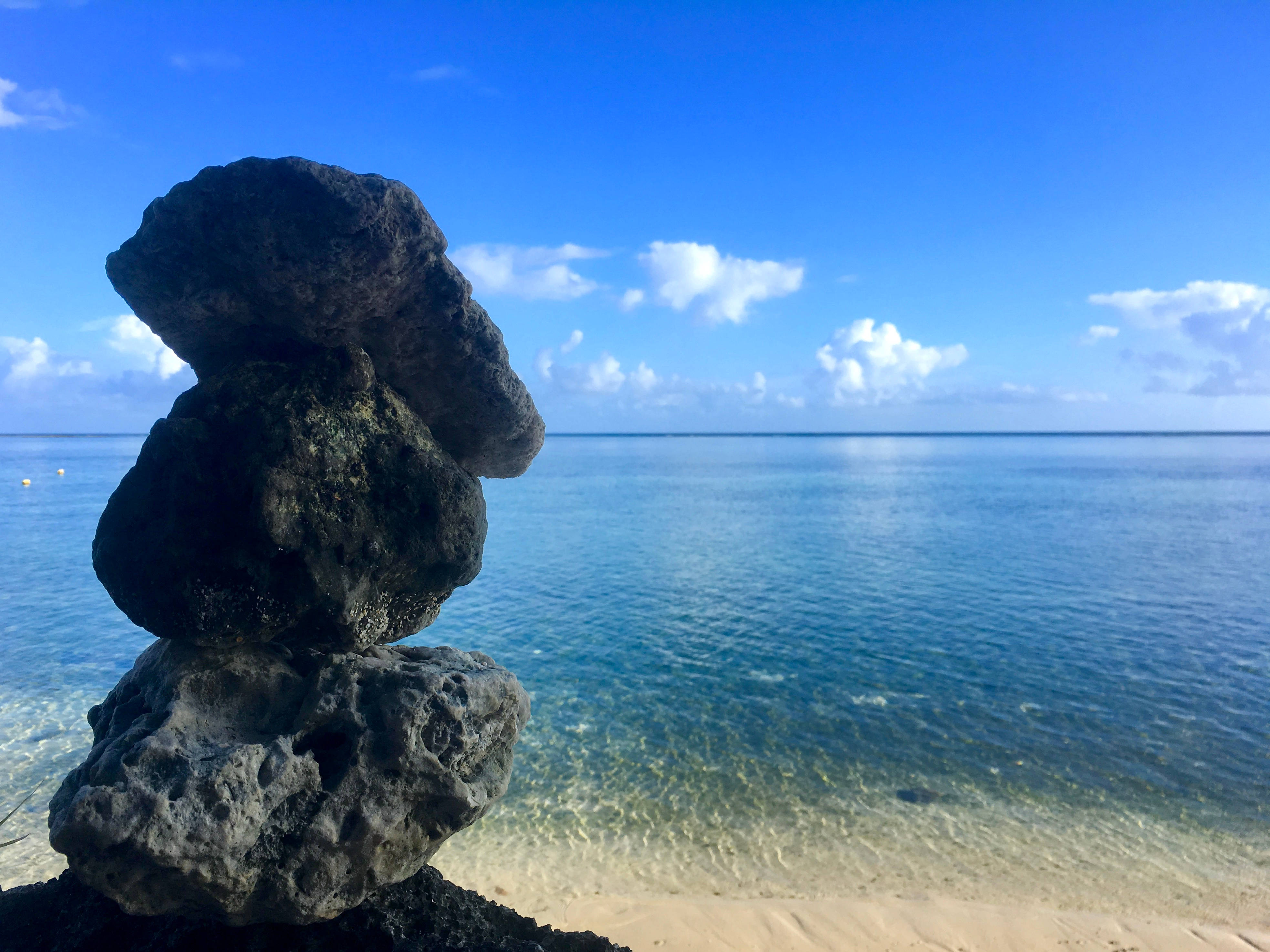 Guam 桌布 壁紙 — 一堆平衡的深色火山岩矗立在沙灘上，清澈的碧綠海水和明亮的藍天點綴著蓬鬆的白雲。