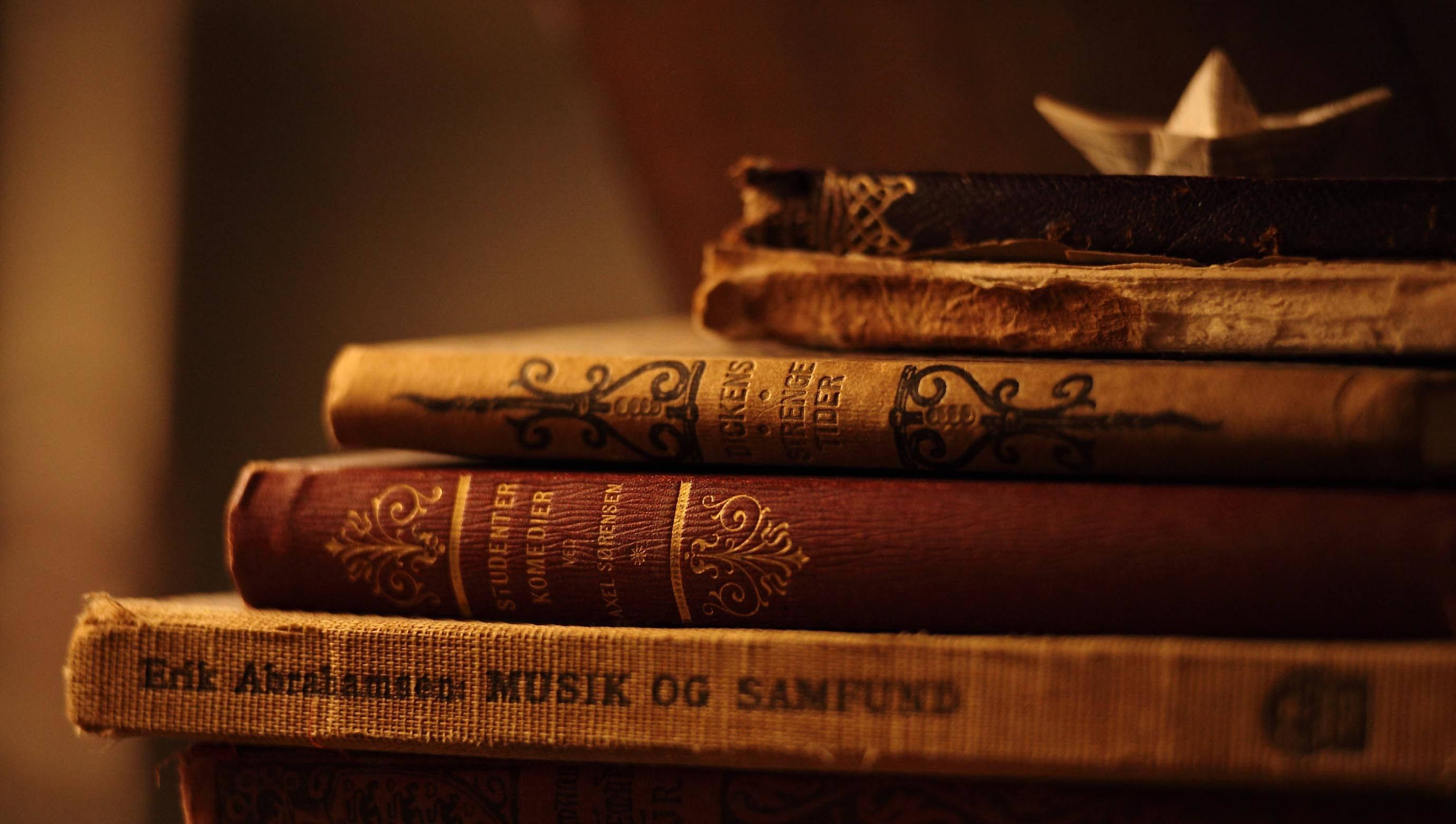 books 桌布 壁紙 — 一堆復古書籍，書脊裝飾華麗，沐浴在溫暖的金色光芒中，頂部放著一艘精緻的紙船。