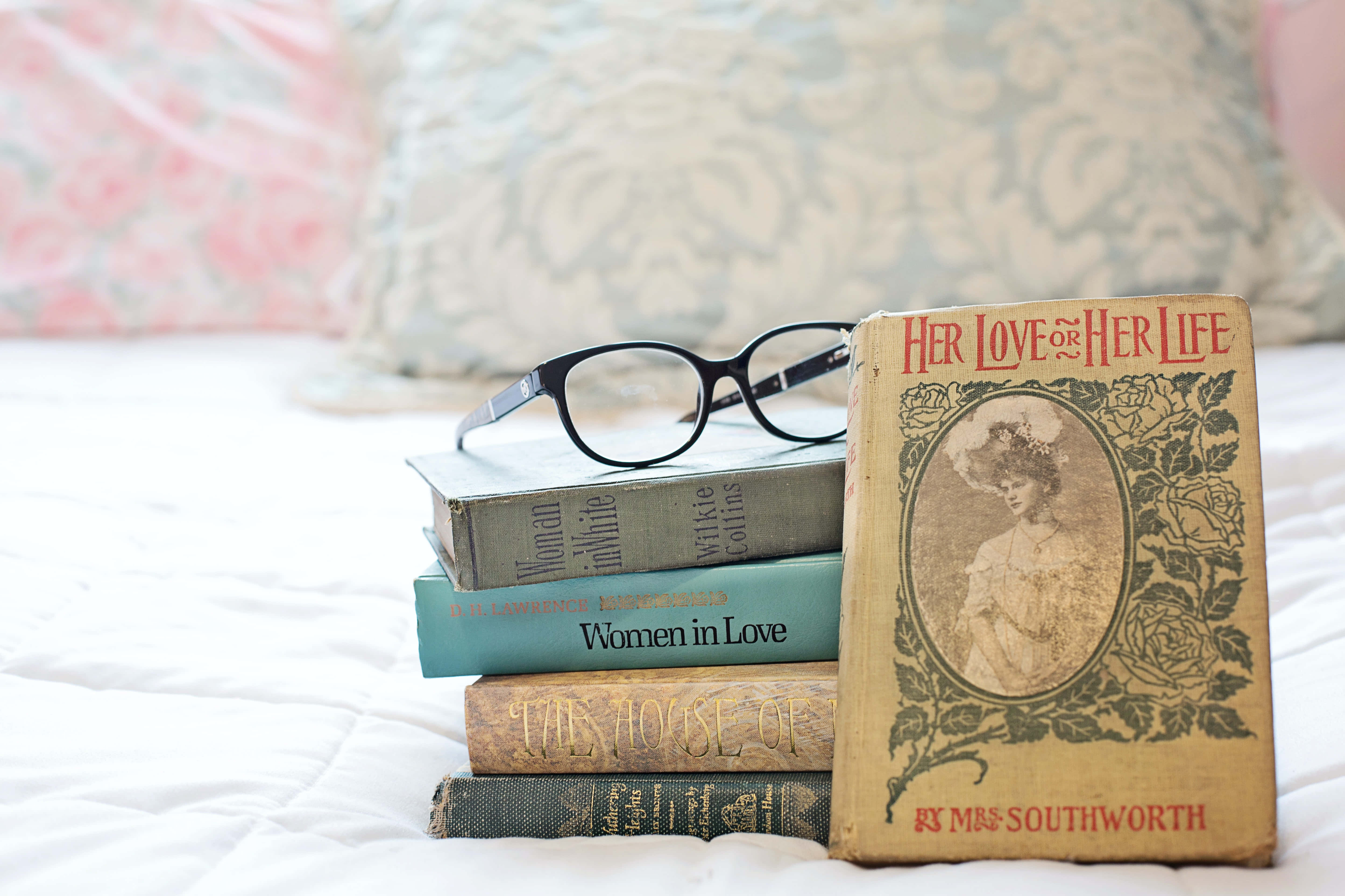 Romance Literature 桌布 壁紙 — 一堆復古書籍配上一副黑框眼鏡，放在白色床罩上，旁邊是圖案枕頭。