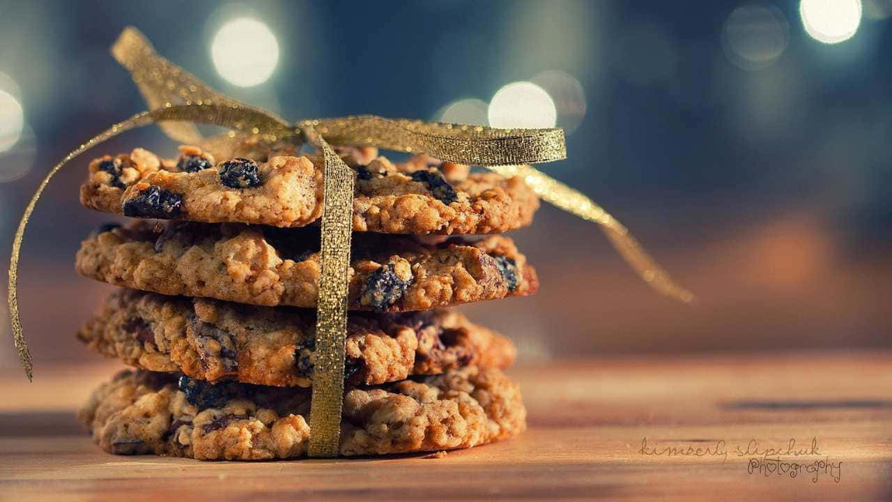 720p Cookies 桌布 壁紙 — 一堆溫暖的燕麥葡萄乾餅乾，用閃閃發光的金色絲帶綁著，放在木質表面上，背景是柔和模糊的散景。