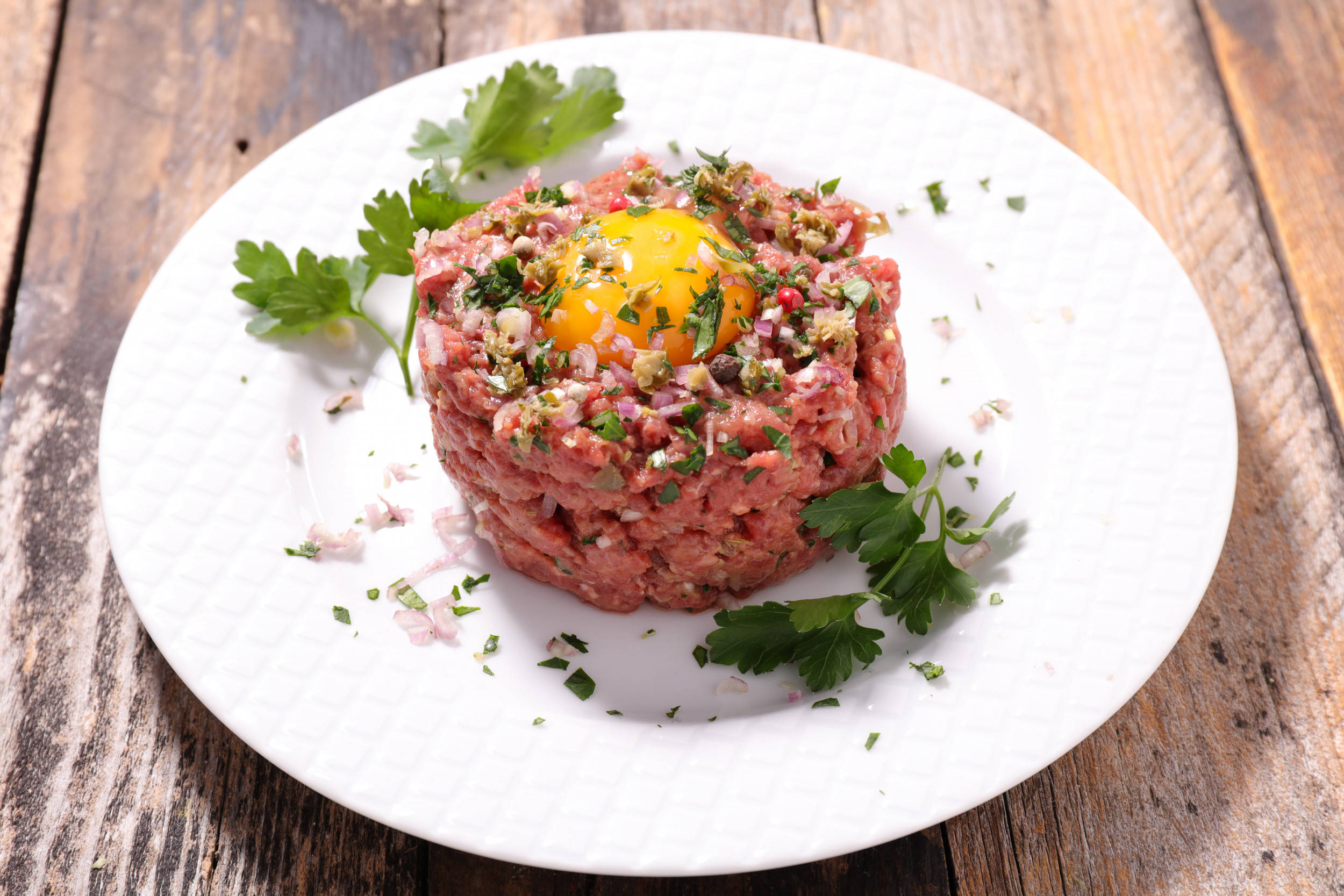 meat 桌布 壁紙 — 一堆生牛肉塔塔，上面放著鮮黃色的生蛋黃和新鮮荷蘭芹，放在白色盤子上，置於質樸的木頭上。