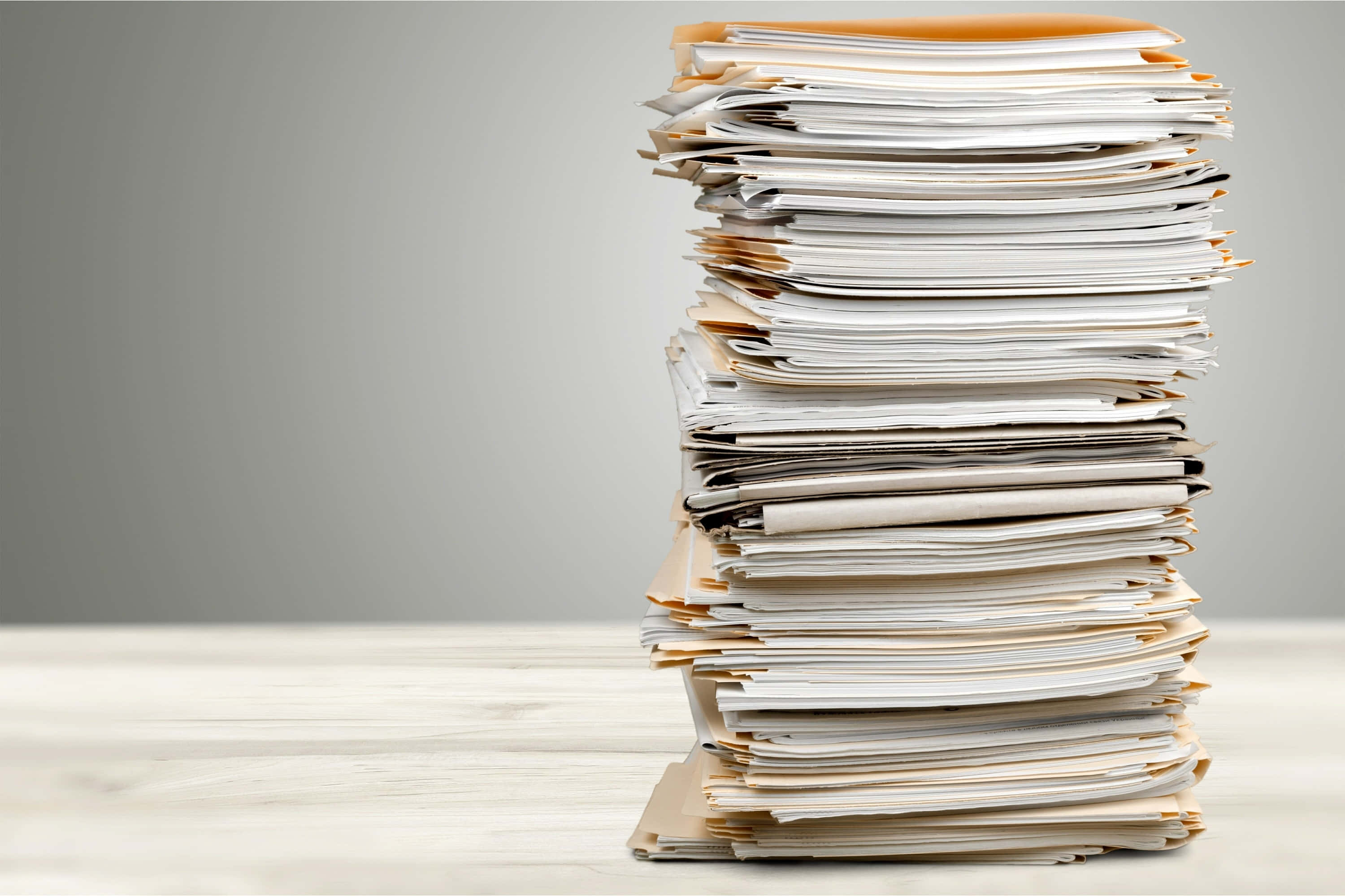 paper 桌布 壁紙 — 一堆白色和米色紙張和信封的高聳雜亂堆疊，在淺色木質表面上不穩地傾斜，背景為中性灰色。