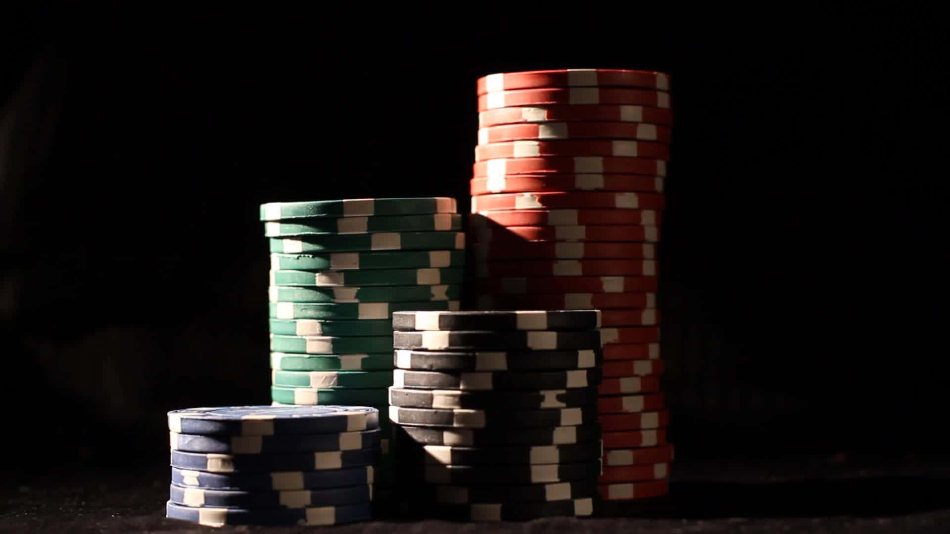 Poker 桌布 壁紙 — 一堆紅色、綠色、黑色和藍色的籌碼，在黑暗背景下被戲劇性的聚光燈照亮。