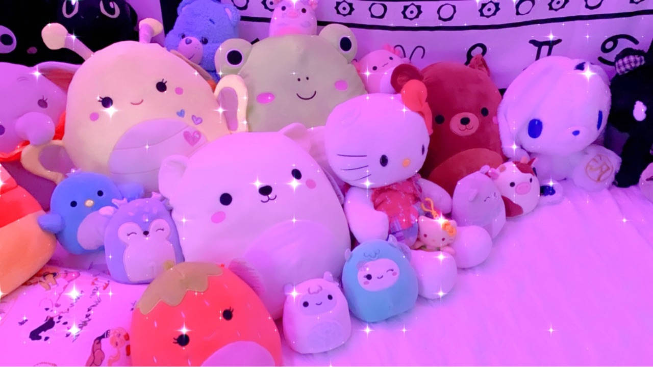 柔軟毛絨玩具在粉紅色燈光下於床上發光的收藏。 — Squishmallows 壁紙