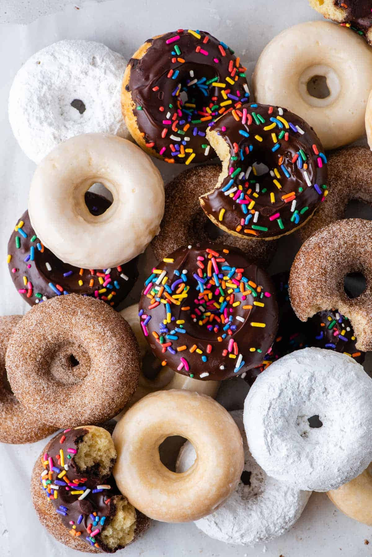Glazed Donut 桌布 壁紙 — 一堆色彩繽紛的綜合甜甜圈，上面有巧克力碎、糖粉和糖霜，擺放在白色表面上。