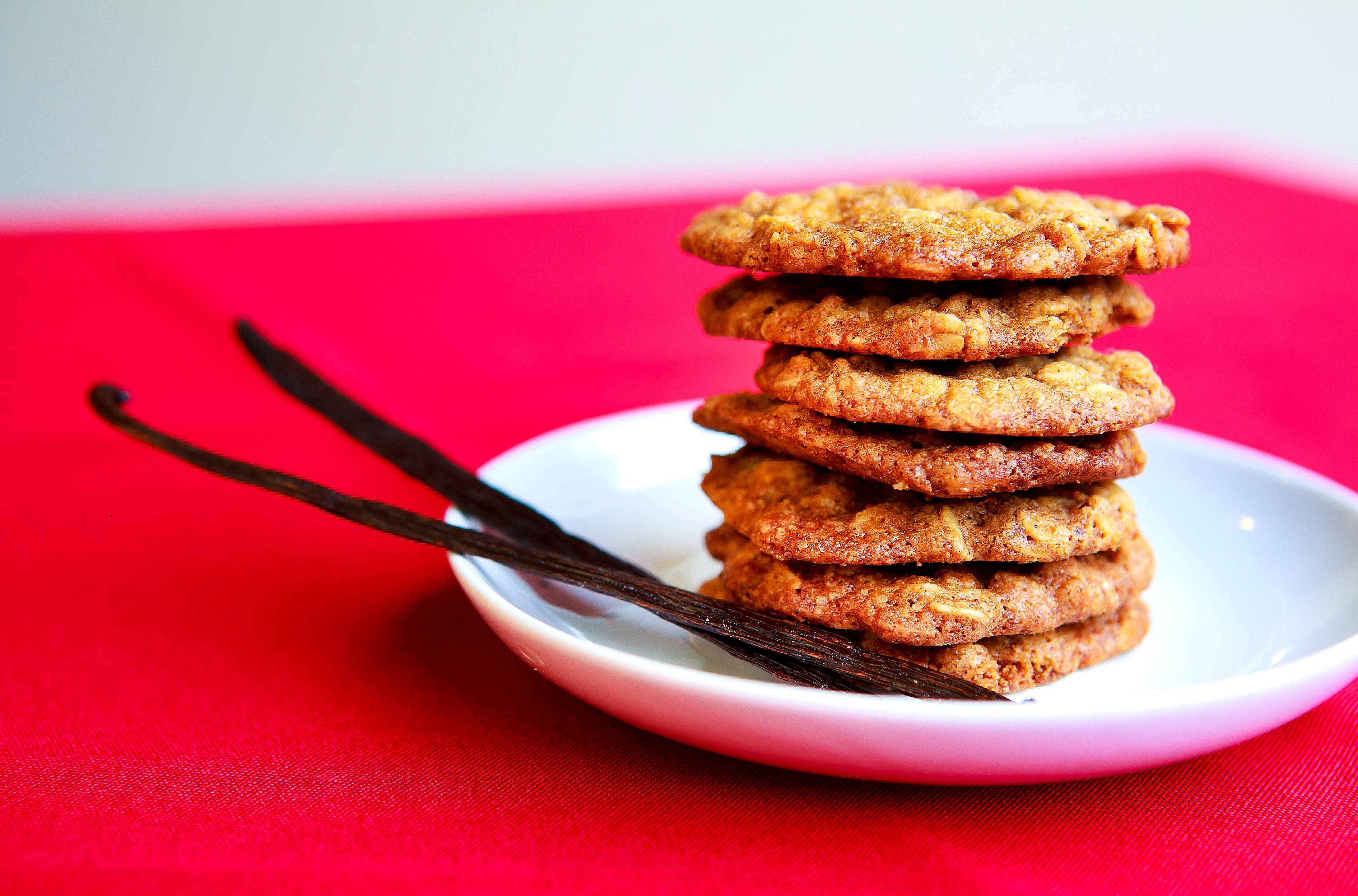 Cookie 桌布 壁紙 — 一堆金黃色的燕麥餅乾放在白色盤子上，旁邊有香草豆，襯托著鮮紅色的布。
