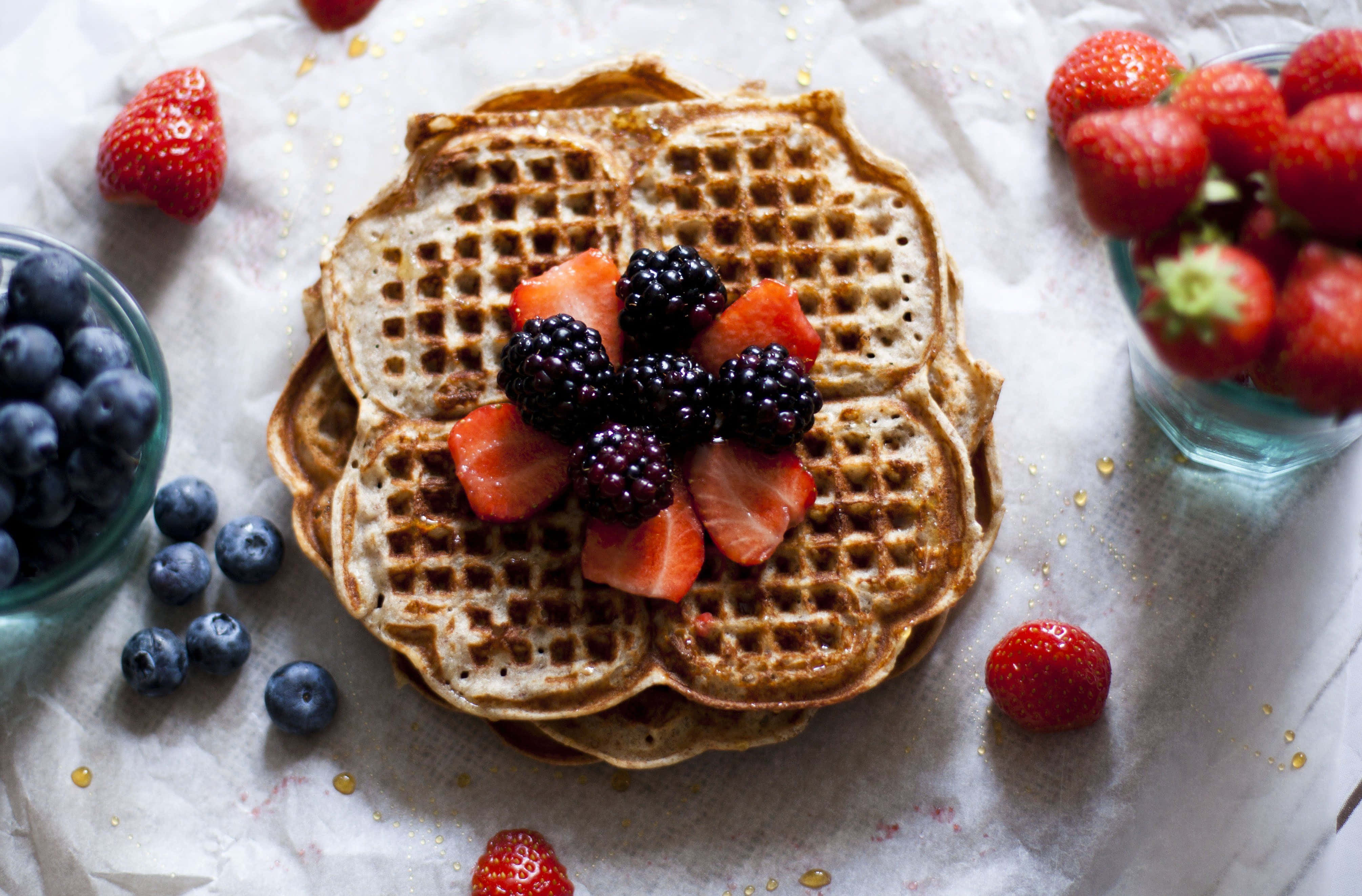 Waffle 桌布 壁紙 — 一堆金黃色鬆餅，上面放上新鮮黑莓和草莓，周圍有藍莓和整顆草莓，放在烘焙紙上。