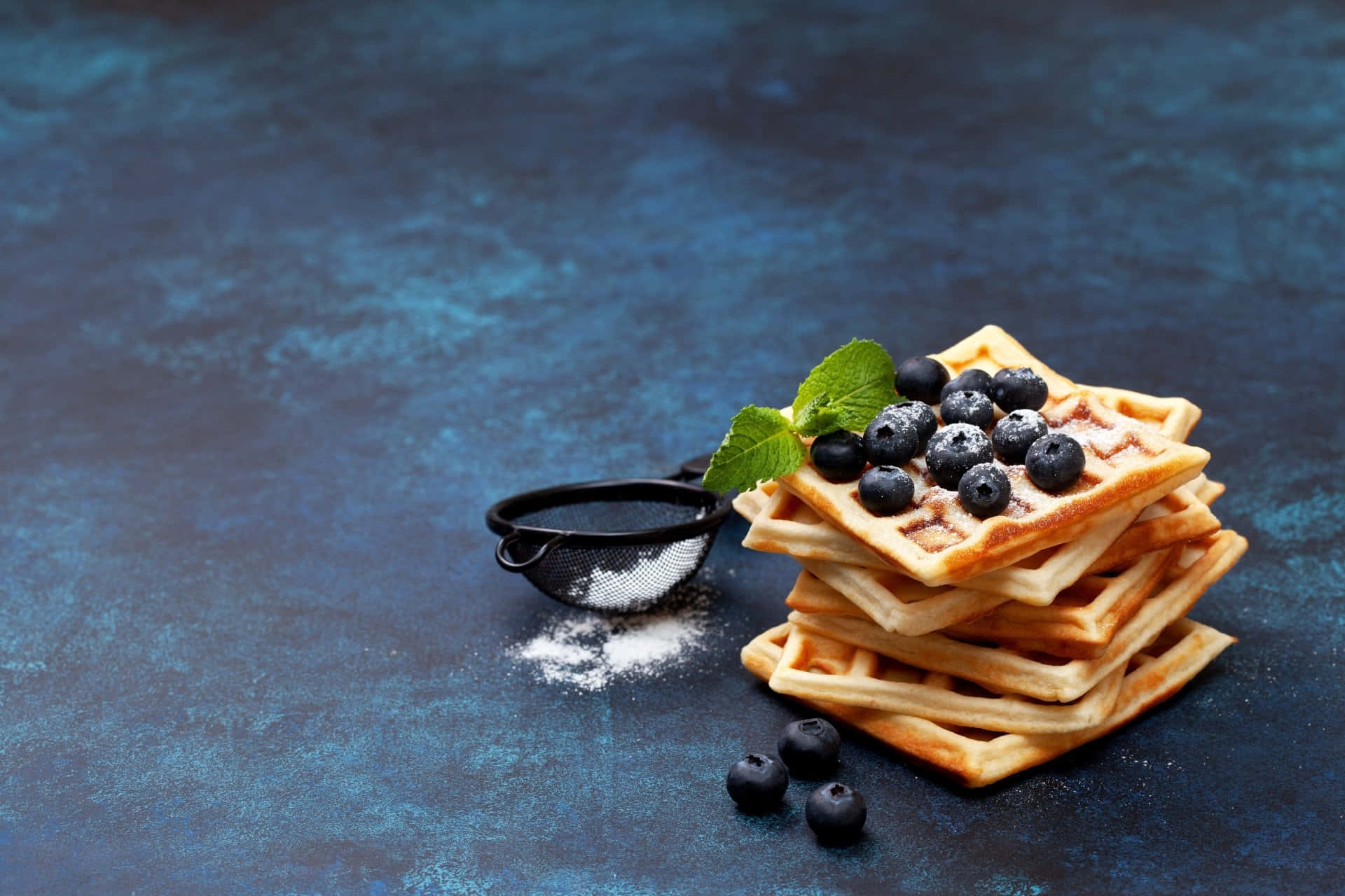 Waffle 桌布 壁紙 — 一堆金黃色鬆餅，上面放有藍莓和薄荷，置於有紋理的藍色表面上，並撒有糖粉。