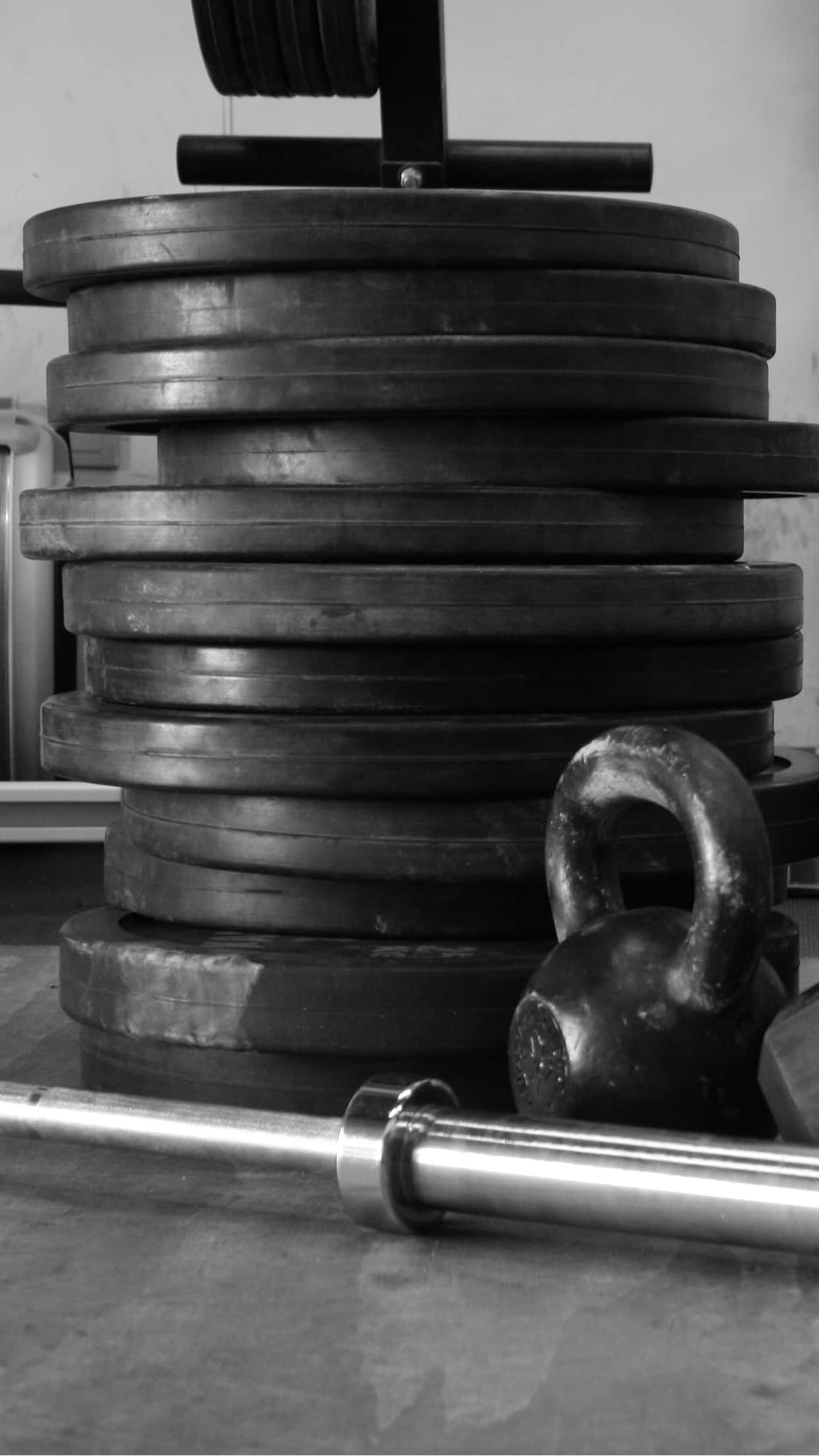 Barbell 桌布 壁紙 — 一堆高疊的槓鈴片和壺鈴放在槓鈴旁邊，身處單色調的健身房環境中。
