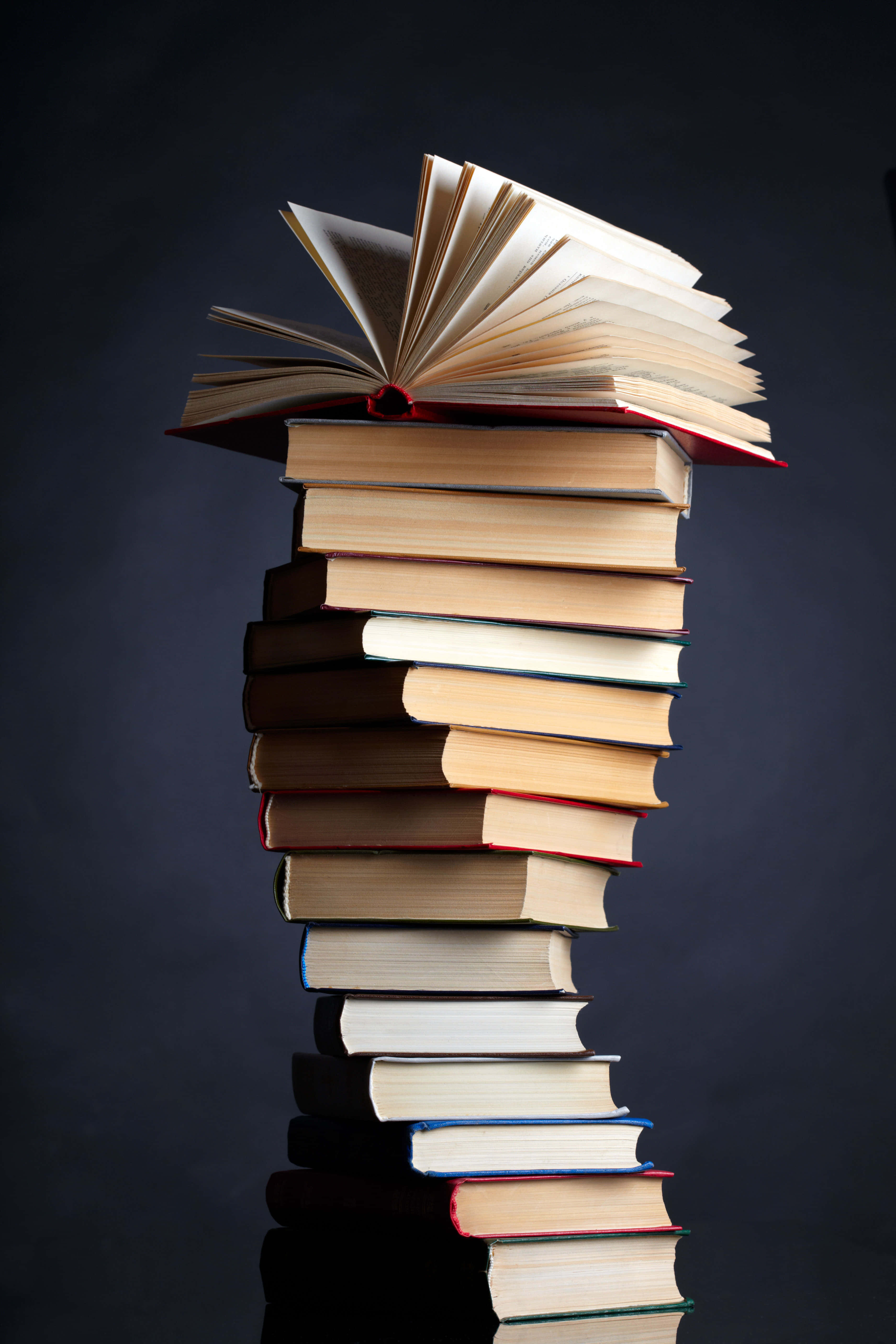 書本 桌布 壁紙 — 一堆高聳且不穩的書，頂部有一本打開的書，背景為深色。