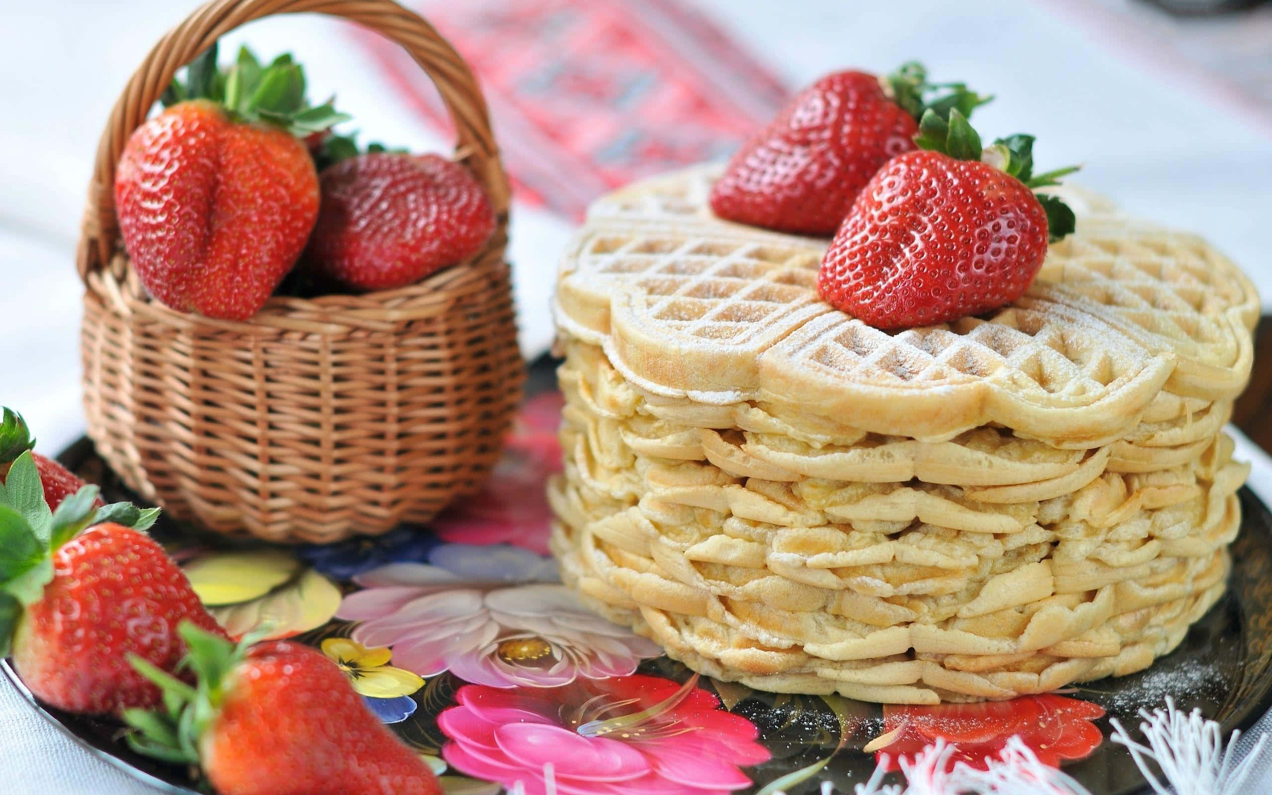 Waffle 桌布 壁紙 — 一堆高高的金黃色鬆餅，撒上糖粉，上面放上新鮮草莓，旁邊有一個編織籃子，裡面有更多草莓，放在一個花卉圖案的盤子上。