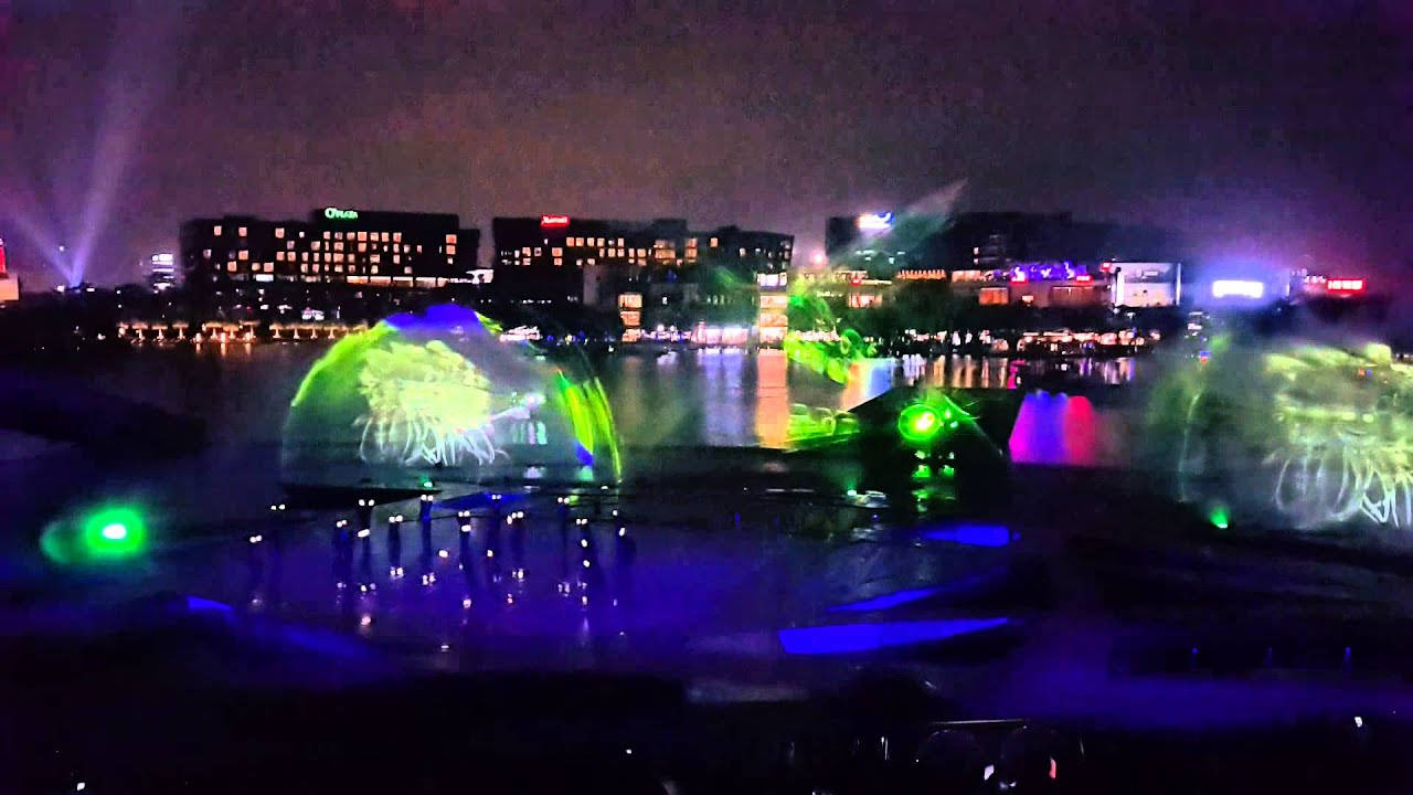 Shenzhen 桌布 壁紙 — 一場令人眼花撩亂的夜間水舞表演，霓虹綠和藍色燈光投射在舞動的噴泉上，映襯著城市天際線。