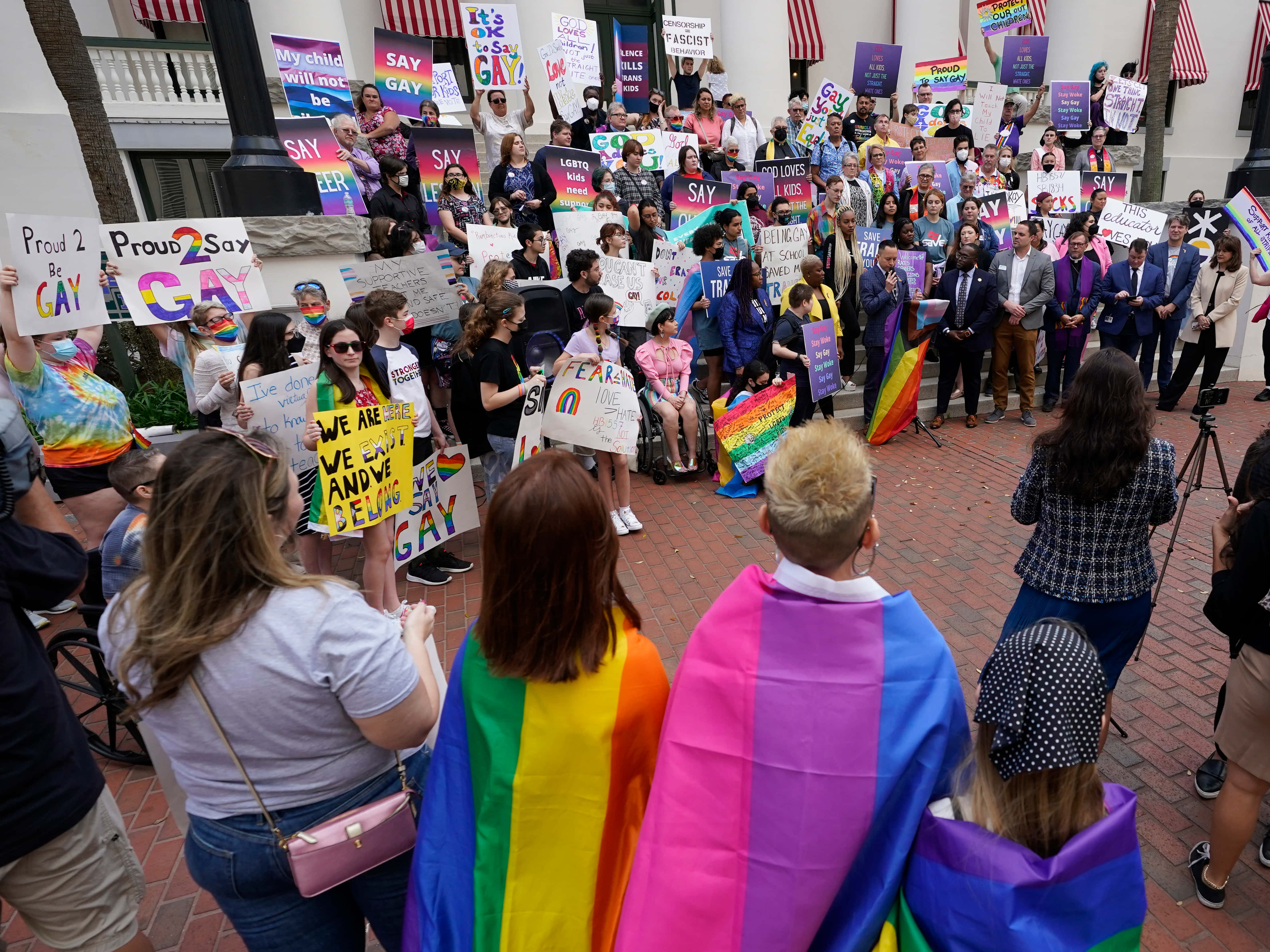 男同志 桌布 壁紙 — 一場充滿活力的 LGBTQ+ 驕傲遊行，多元參與者在磚砌廣場上於日光下舉著彩虹標語和旗幟。