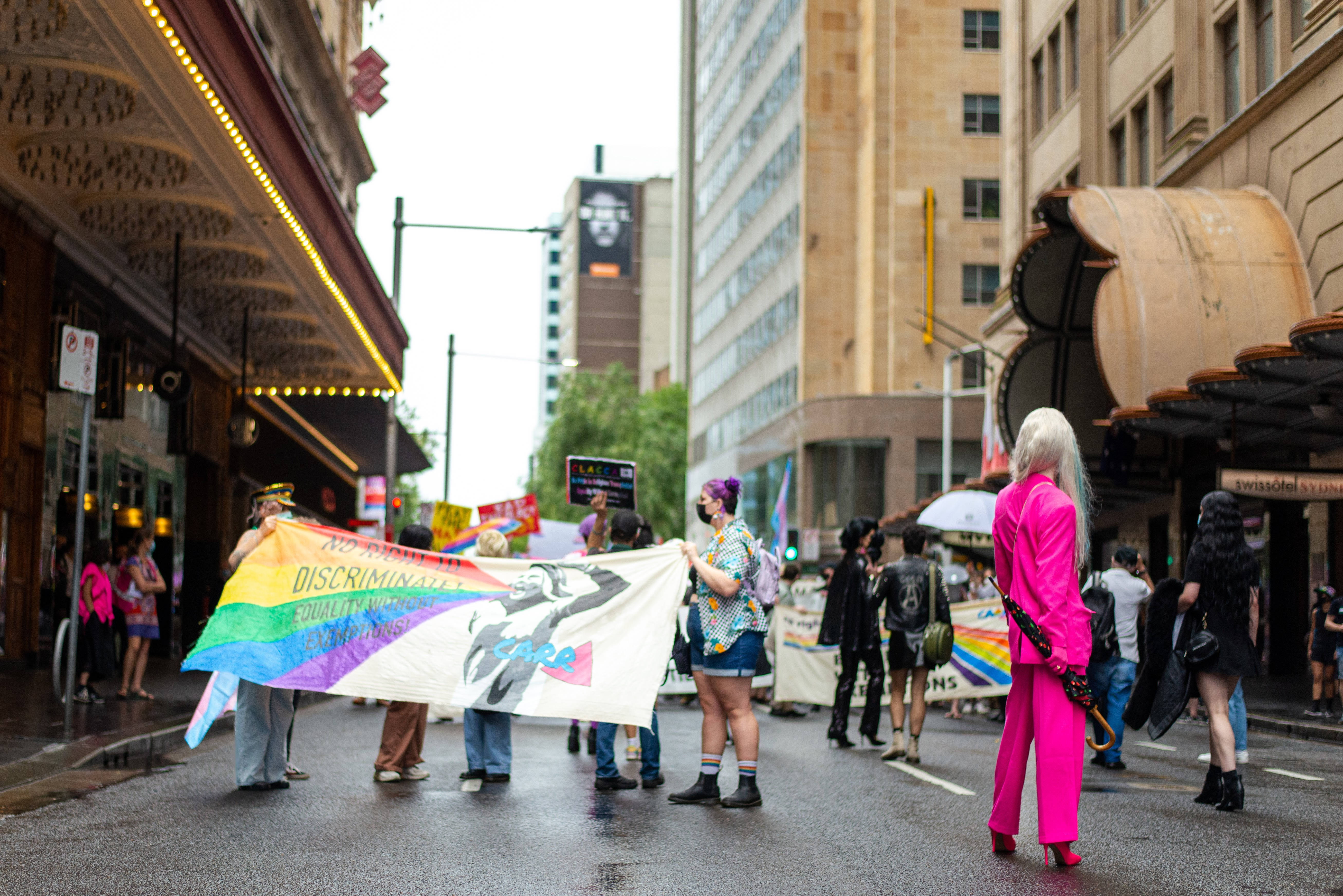 Binary 桌布 壁紙 — 一場充滿活力的LGBTQ+驕傲遊行在濕漉漉的城市街道上展開，參與者高舉彩虹旗幟和標語，在多雲的天空下要求平等。