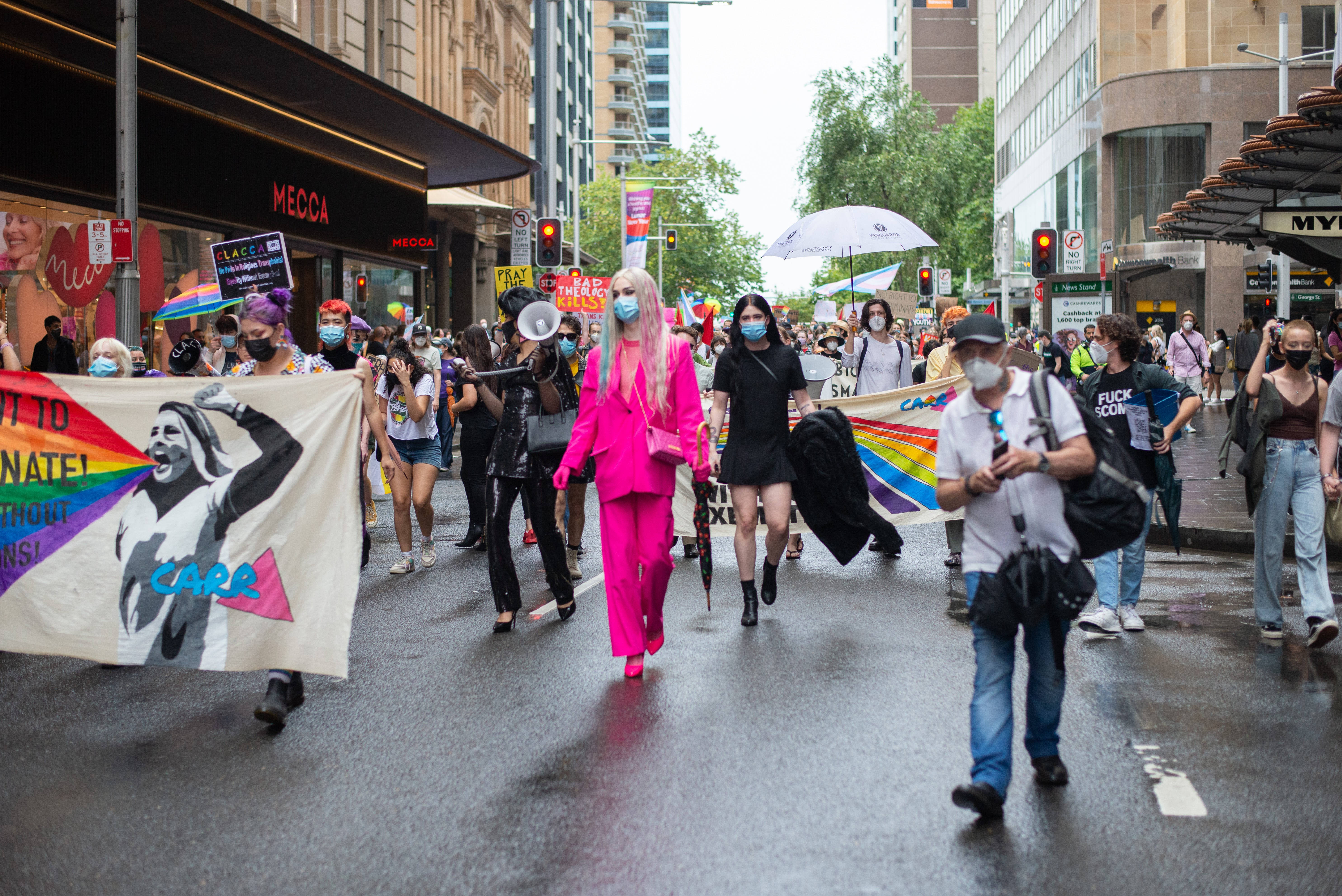 Binary 桌布 壁紙 — 一場充滿活力的LGBTQ+驕傲遊行沿著濕漉漉的城市街道前進，彩虹旗飄揚，抗議者戴著口罩和穿著色彩繽紛的服裝。