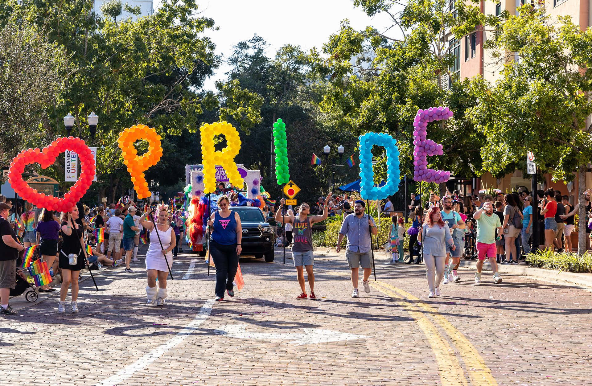 驕傲 桌布 壁紙 — 一場充滿活力的Pride遊行沿著綠樹成蔭的街道前進，參與者手持巨大的彩虹氣球字母拼出PRIDE和一個心形。