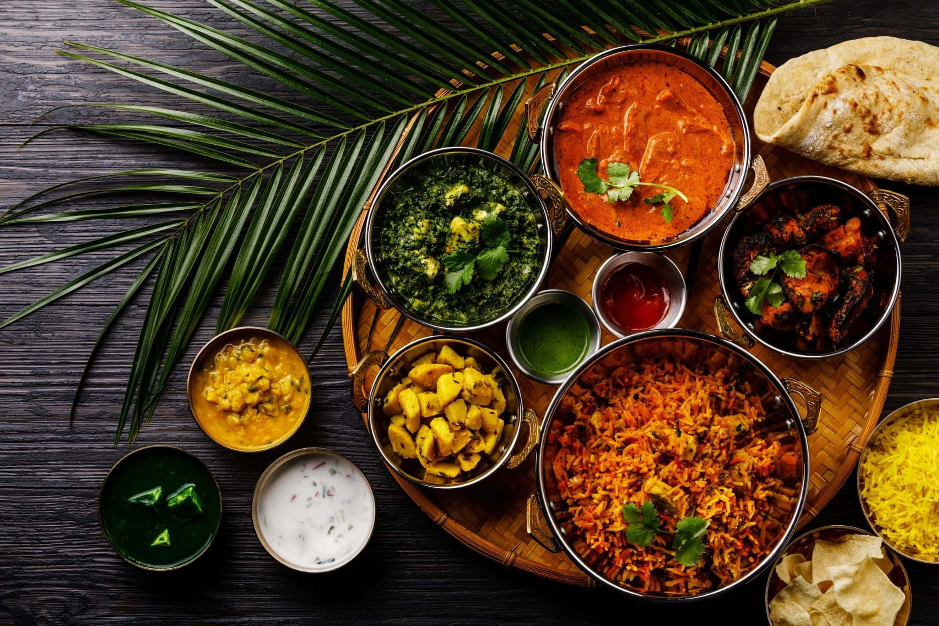 Indian 桌布 壁紙 — 一場充滿活力的印度盛宴，色彩繽紛的咖哩、米飯和烤餅整齊地擺放在木托盤上，旁邊襯有棕櫚葉。