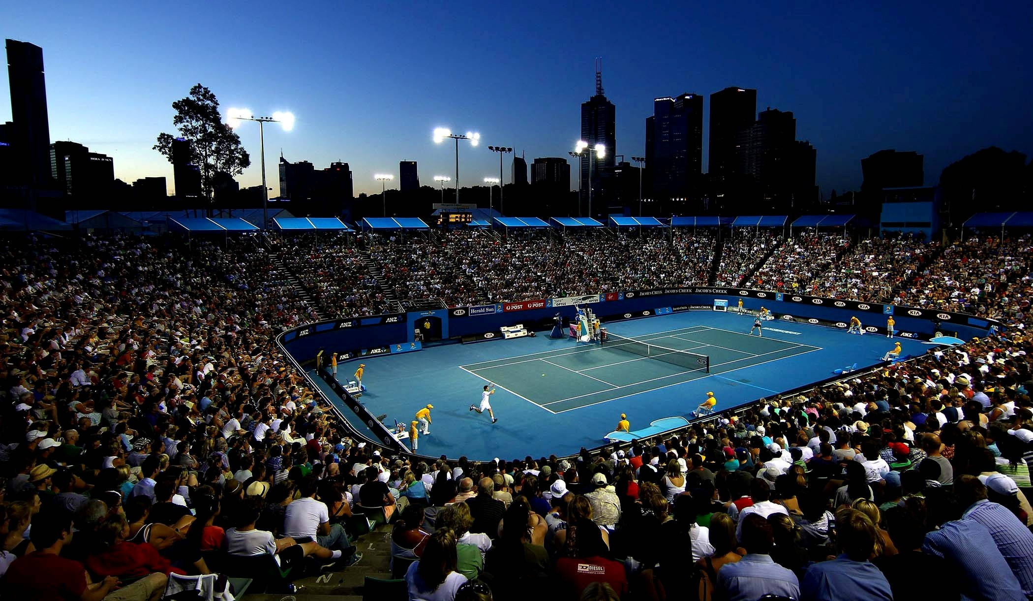 Tennis 桌布 壁紙 — 一場充滿活力的夜間網球比賽在體育場燈光下展開，觀眾席座無虛席，背景是城市天際線。