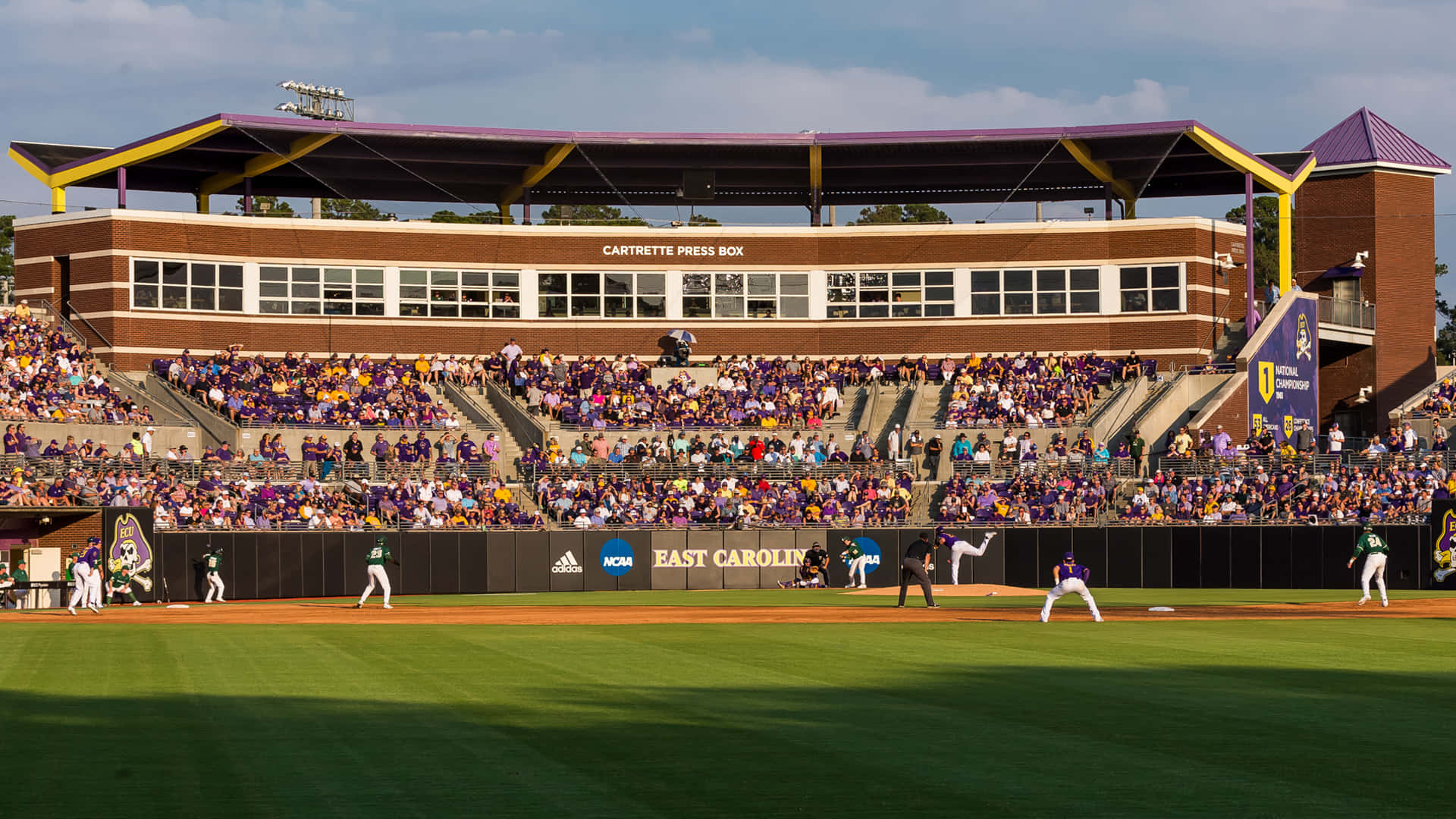 College Baseball 桌布 壁紙 — 一場充滿活力的大學棒球比賽正在進行，天空多雲，身穿紫色和金色服裝的球迷擠滿了看台。