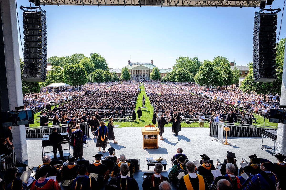 Graduation 桌布 壁紙 — 一場充滿活力的戶外畢業典禮，數千名學生身穿畢業袍和學士帽，坐在藍天白雲下的草地上。