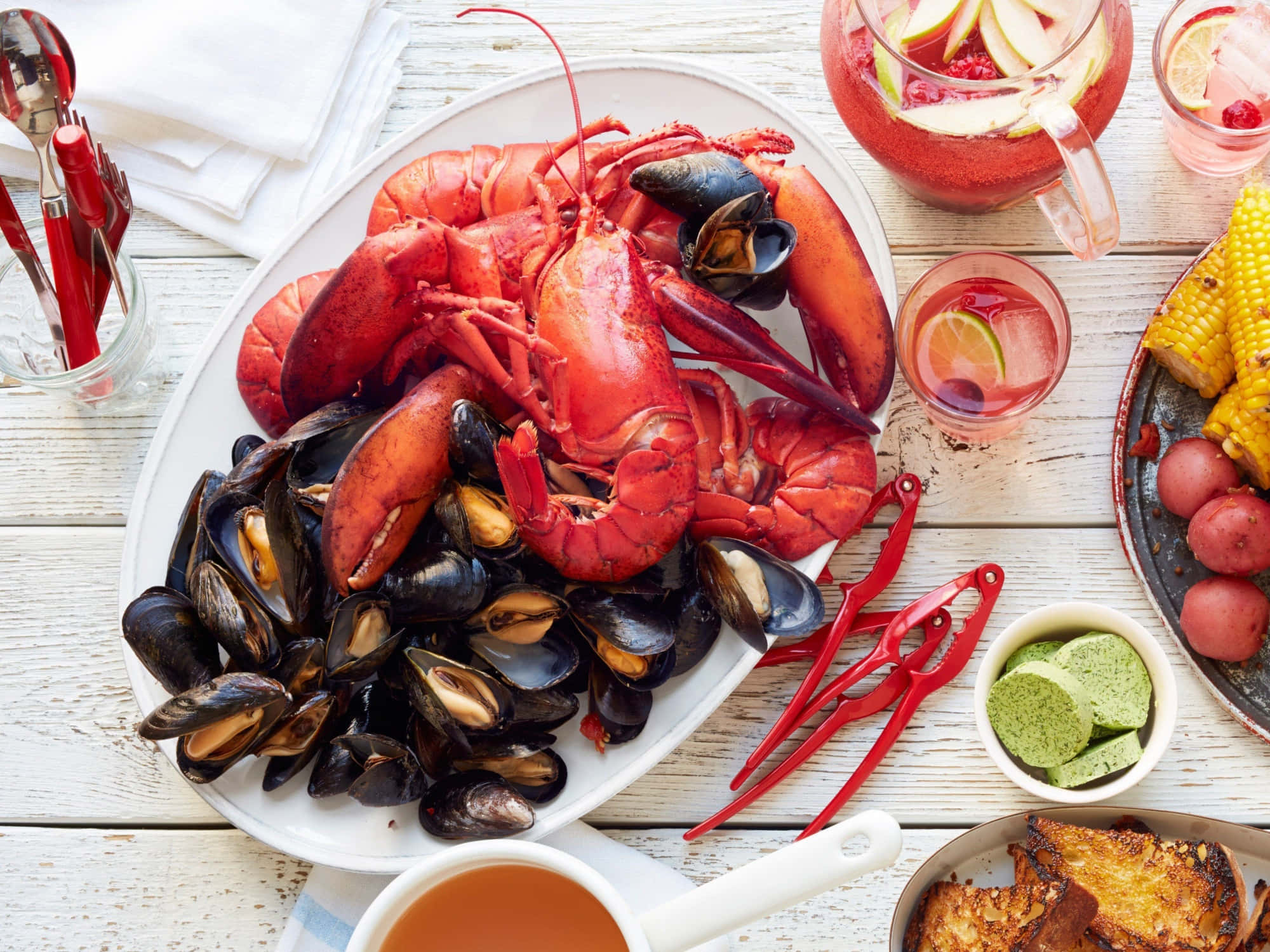 Seafood 桌布 壁紙 — 一場充滿活力的海鮮盛宴，有鮮紅色的龍蝦和黑色淡菜放在白色盤子上，周圍有玉米、馬鈴薯和清涼飲料，放在鄉村風格的木桌上。