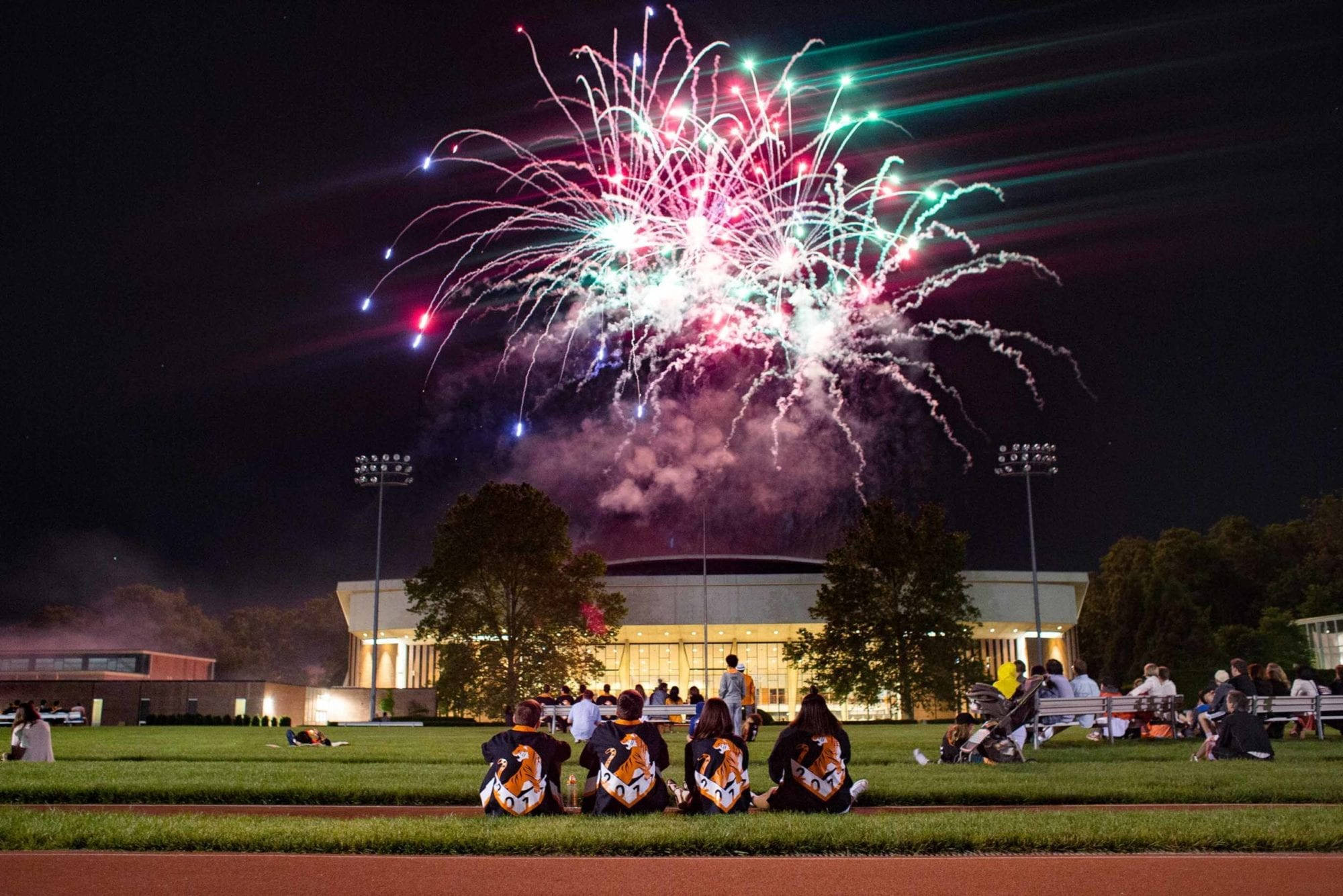 Fireworks 桌布 壁紙 — 一場充滿活力的煙火表演在夜間體育場上空綻放，觀眾坐在草地上觀看。