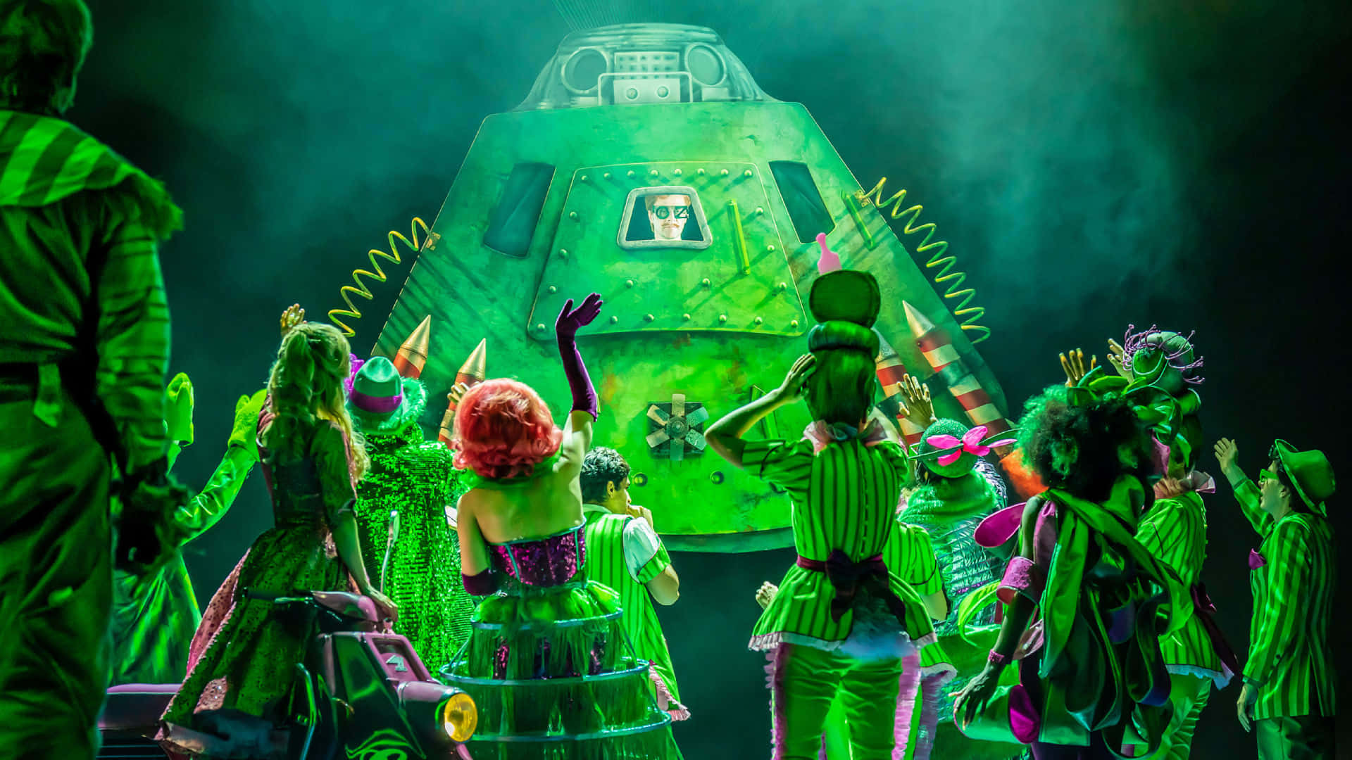 wizard of oz 桌布 壁紙 — 一場充滿活力的舞台表演，色彩繽紛的表演者聚集在一個巨大的綠色太空船周圍，在戲劇性的綠色燈光下。