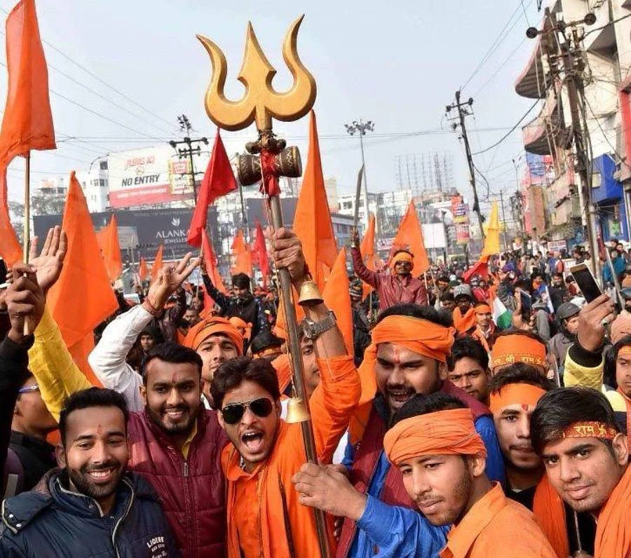 Indian 桌布 壁紙 — 一場充滿活力的街頭抗議，戴著橙色頭巾的男子在晴朗的天空下揮舞旗幟並手持金色三叉戟標誌。