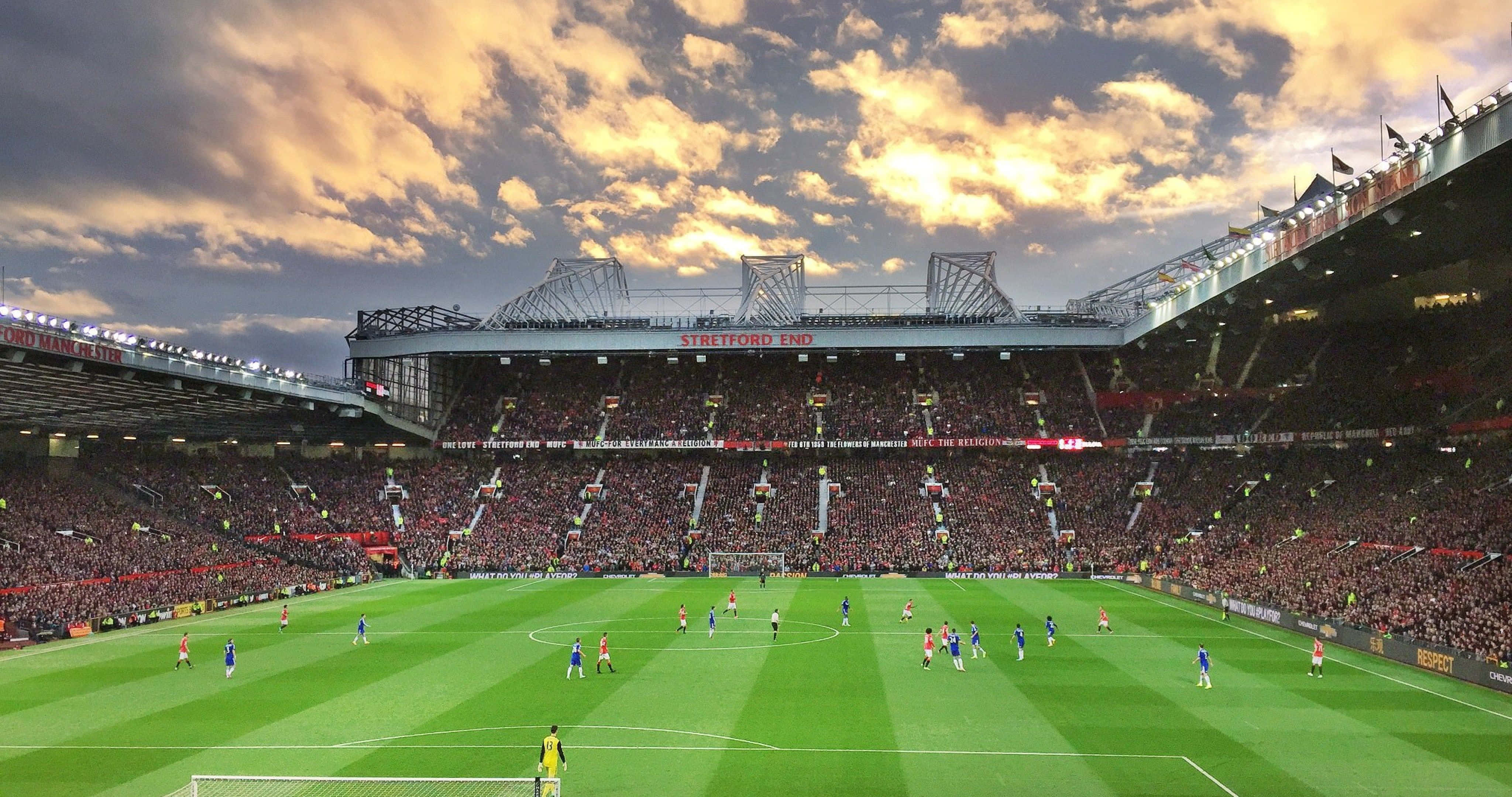Soccer 4K 桌布 壁紙 — 一場充滿活力的足球比賽在奧脫福球場上演，戲劇性的日落天空下，球場內擠滿了歡呼的球迷。
