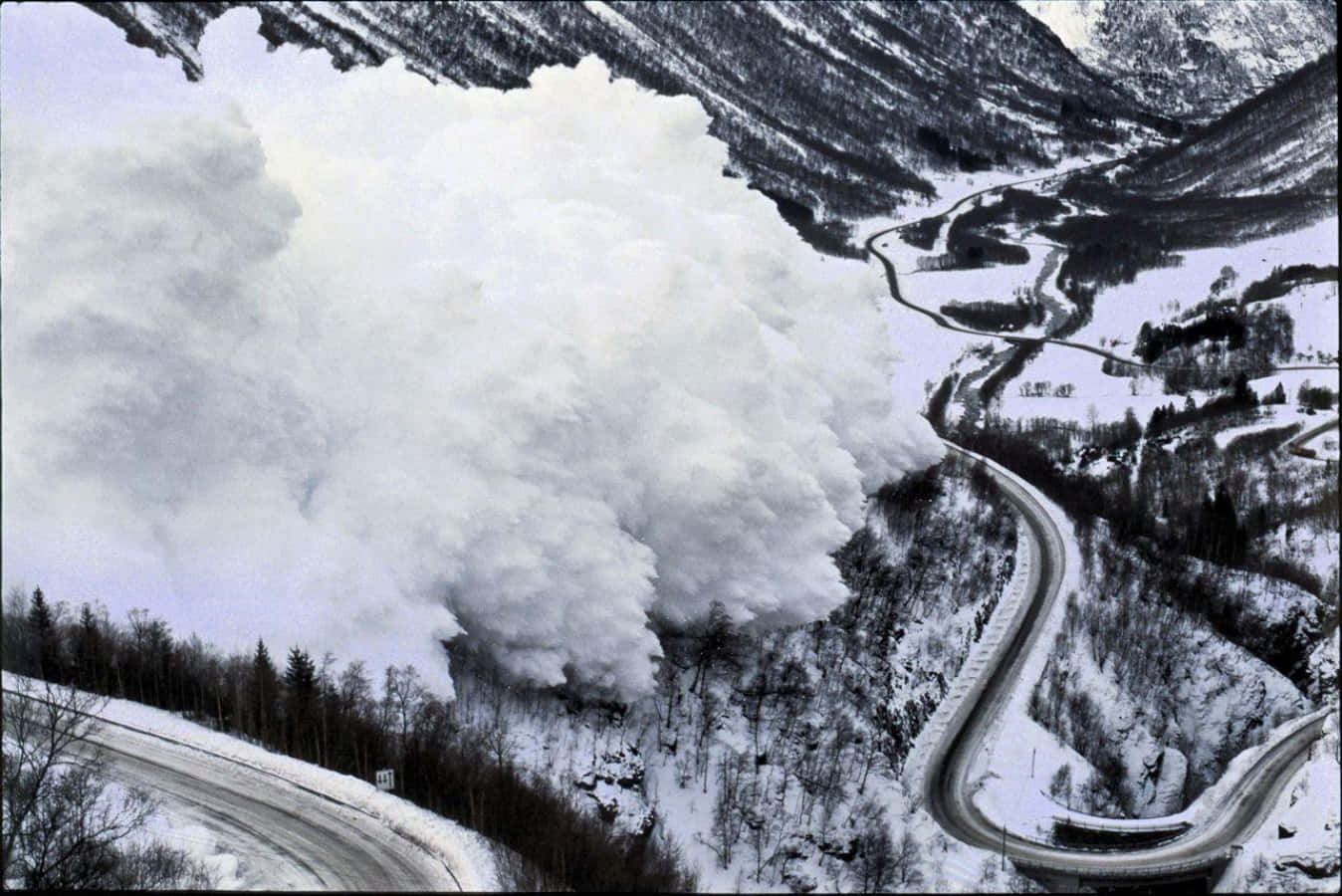 Natural Disaster 桌布 壁紙 — 一場巨大的雪崩從白雪皚皚的山坡上傾瀉而下，將下方蜿蜒的道路掩埋在冬季仙境中。