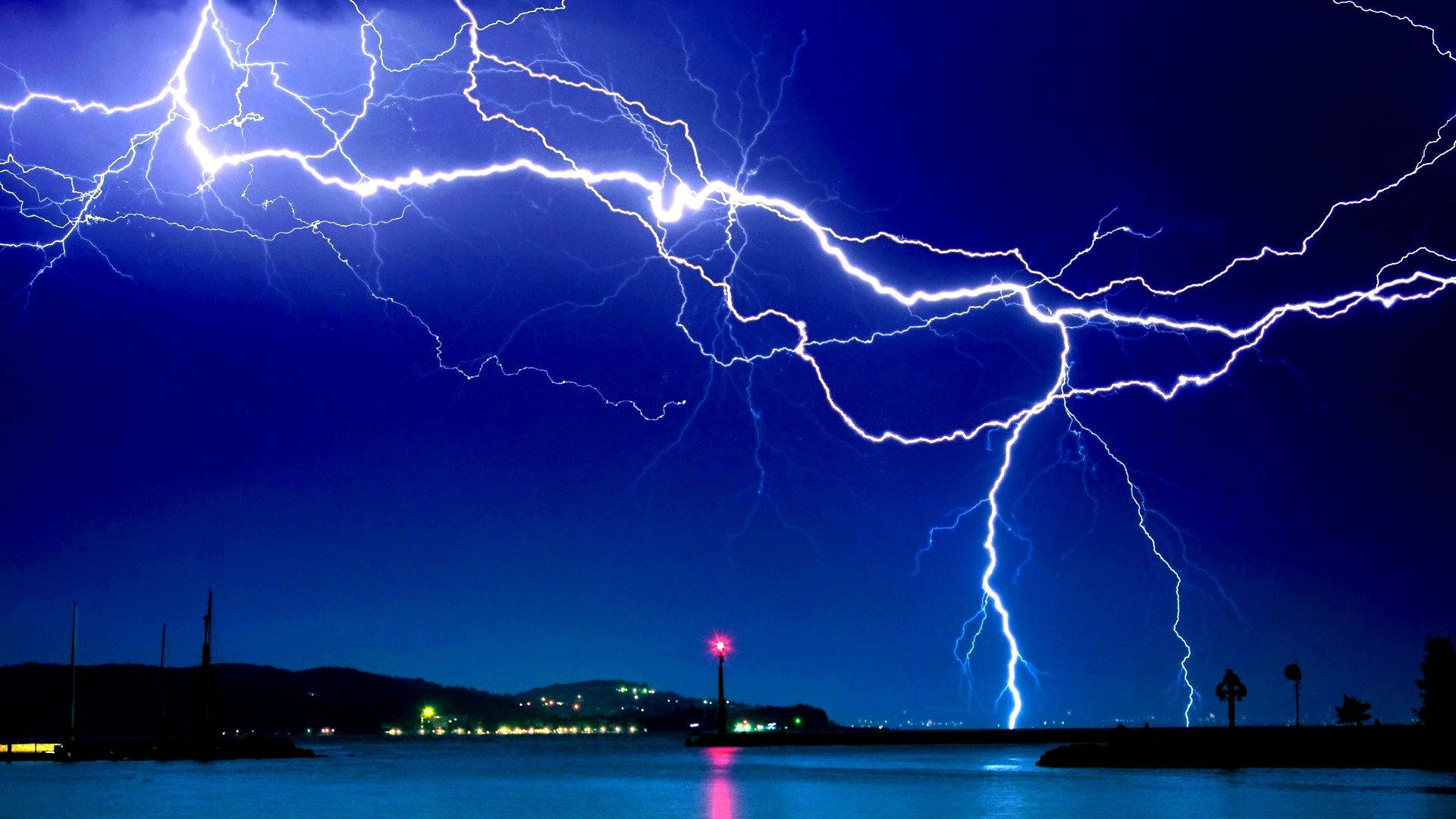 閃電 桌布 壁紙 — 一場戲劇性的閃電風暴照亮了平靜水域上空深藍色的夜空，遠處有城市燈光和燈塔。