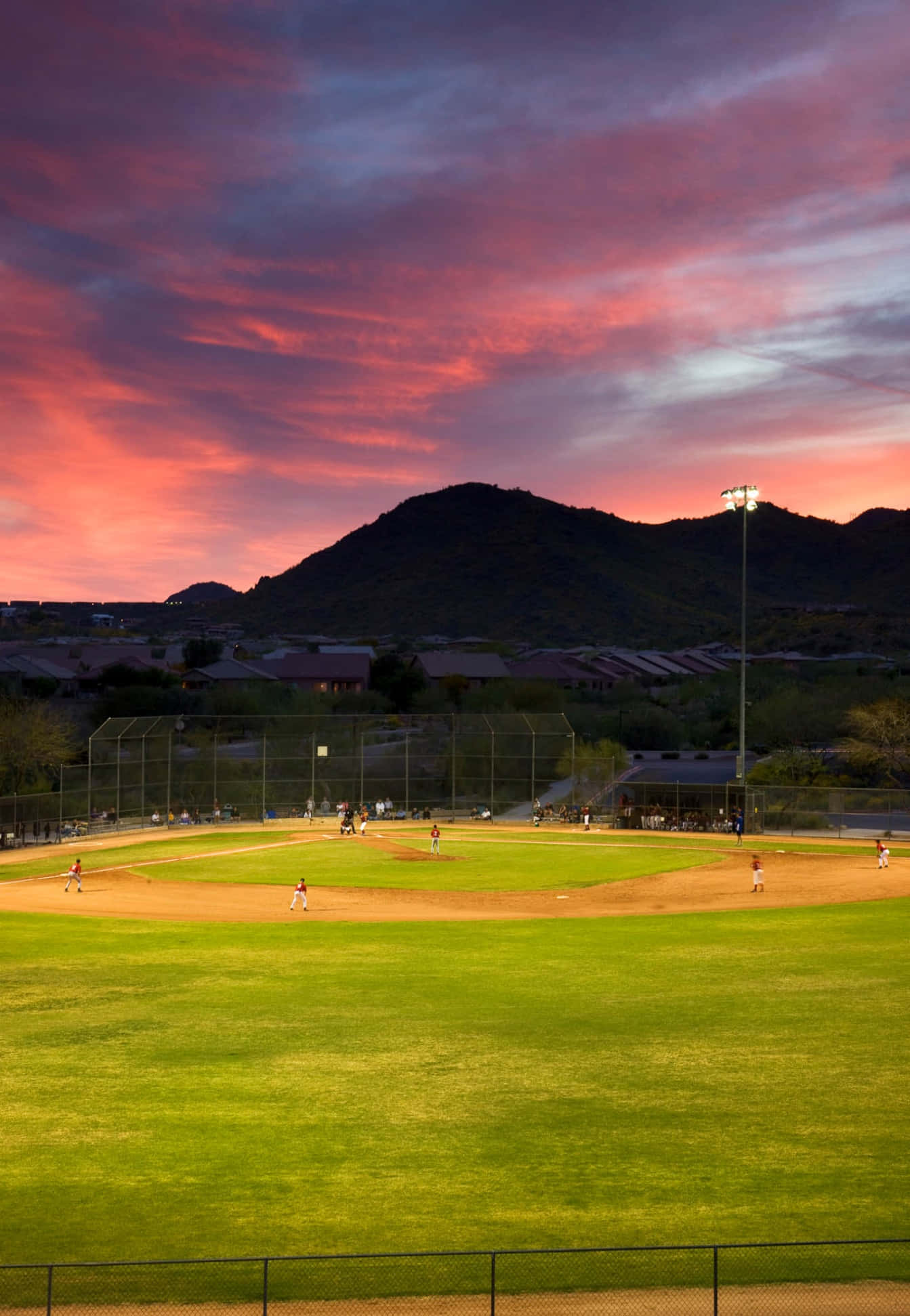 Baseball Field 桌布 壁紙 — 一場棒球比賽在戲劇性的粉紅和紫色日落天空下展開，背景是輪廓分明的山脈。