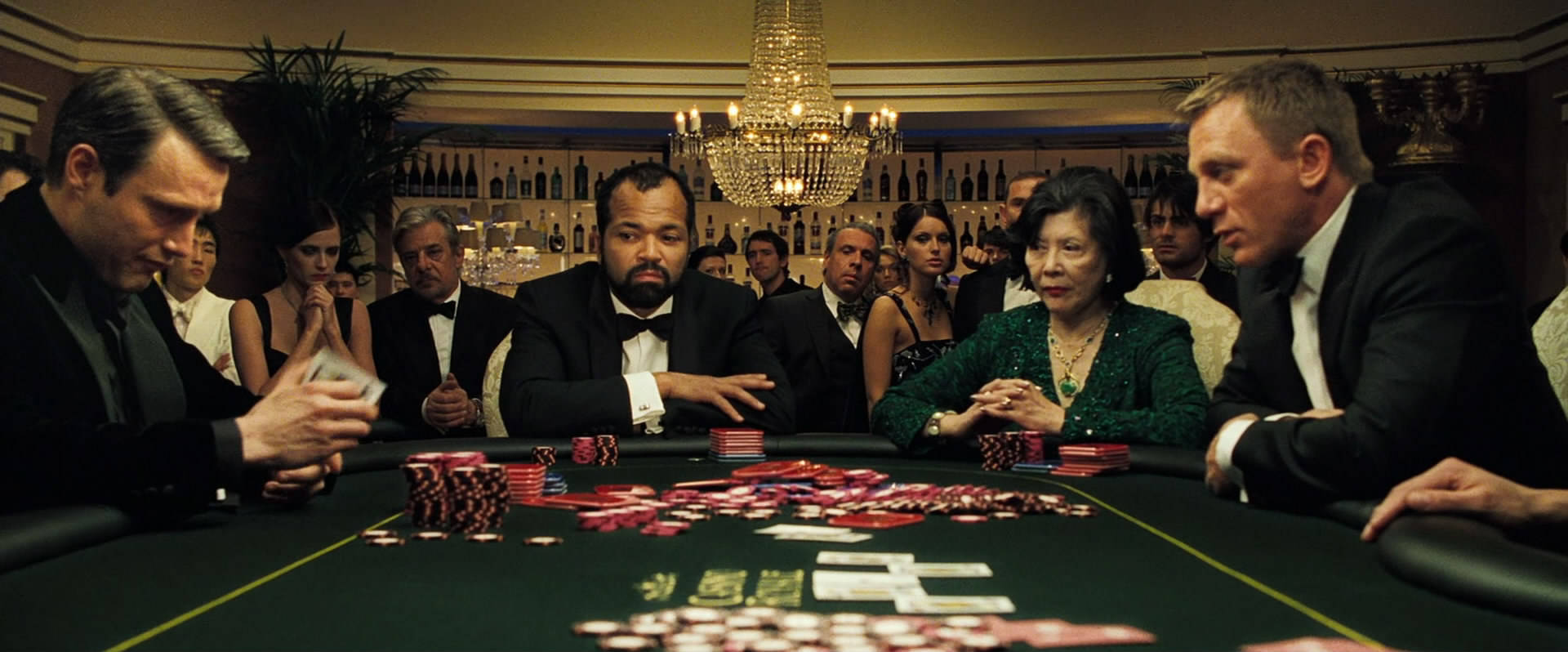 Poker 桌布 壁紙 — 一場激烈的撲克遊戲在豪華賭場中展開，玩家身穿燕尾服，周圍是閃爍的水晶吊燈和成堆的紅色籌碼。