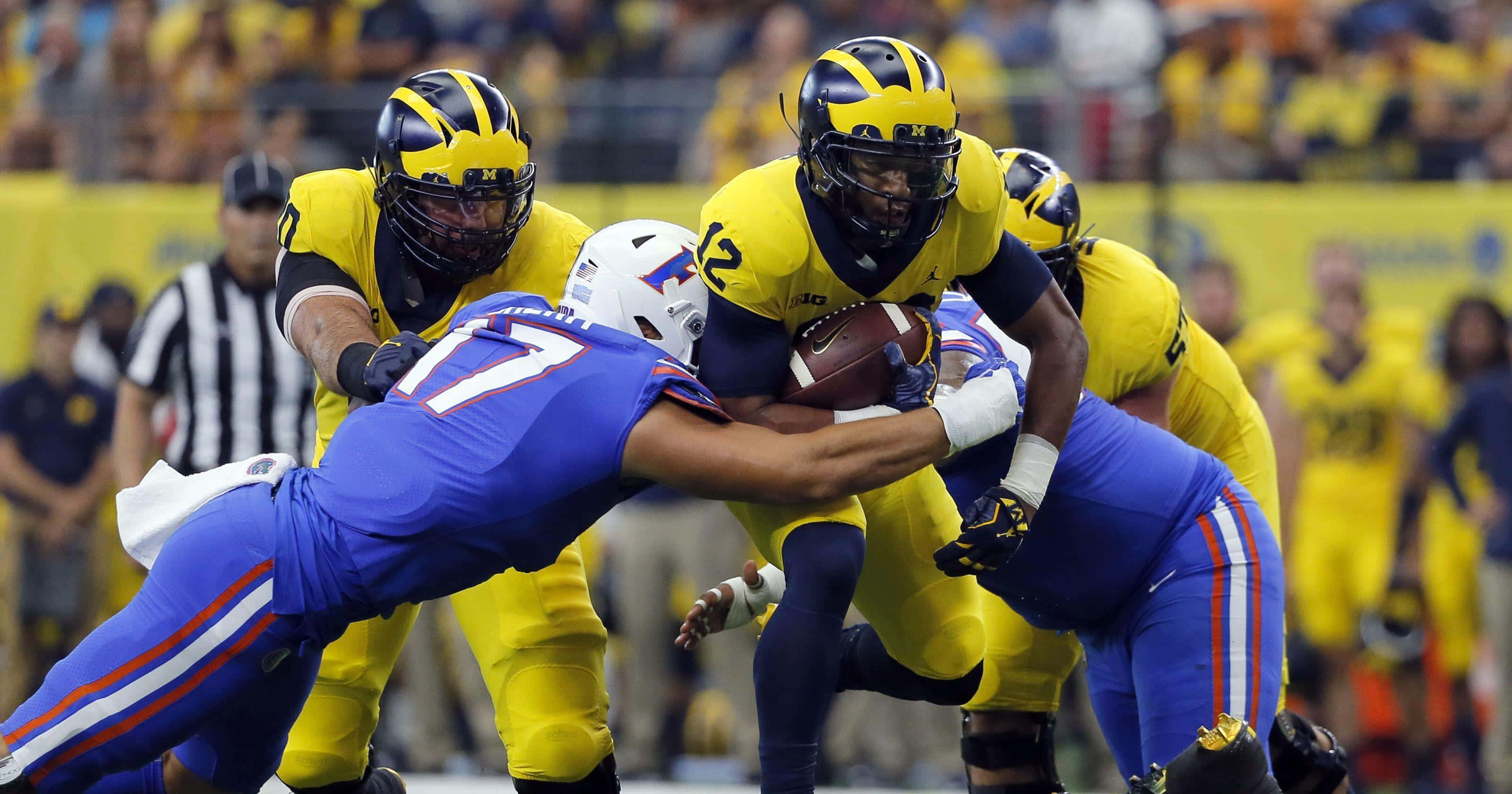 Michigan Football 桌布 壁紙 — 一場激烈的比賽中，密西根州足球員身穿黃色球衣緊抱球，被佛羅里達州藍色球衣球員擒抱。