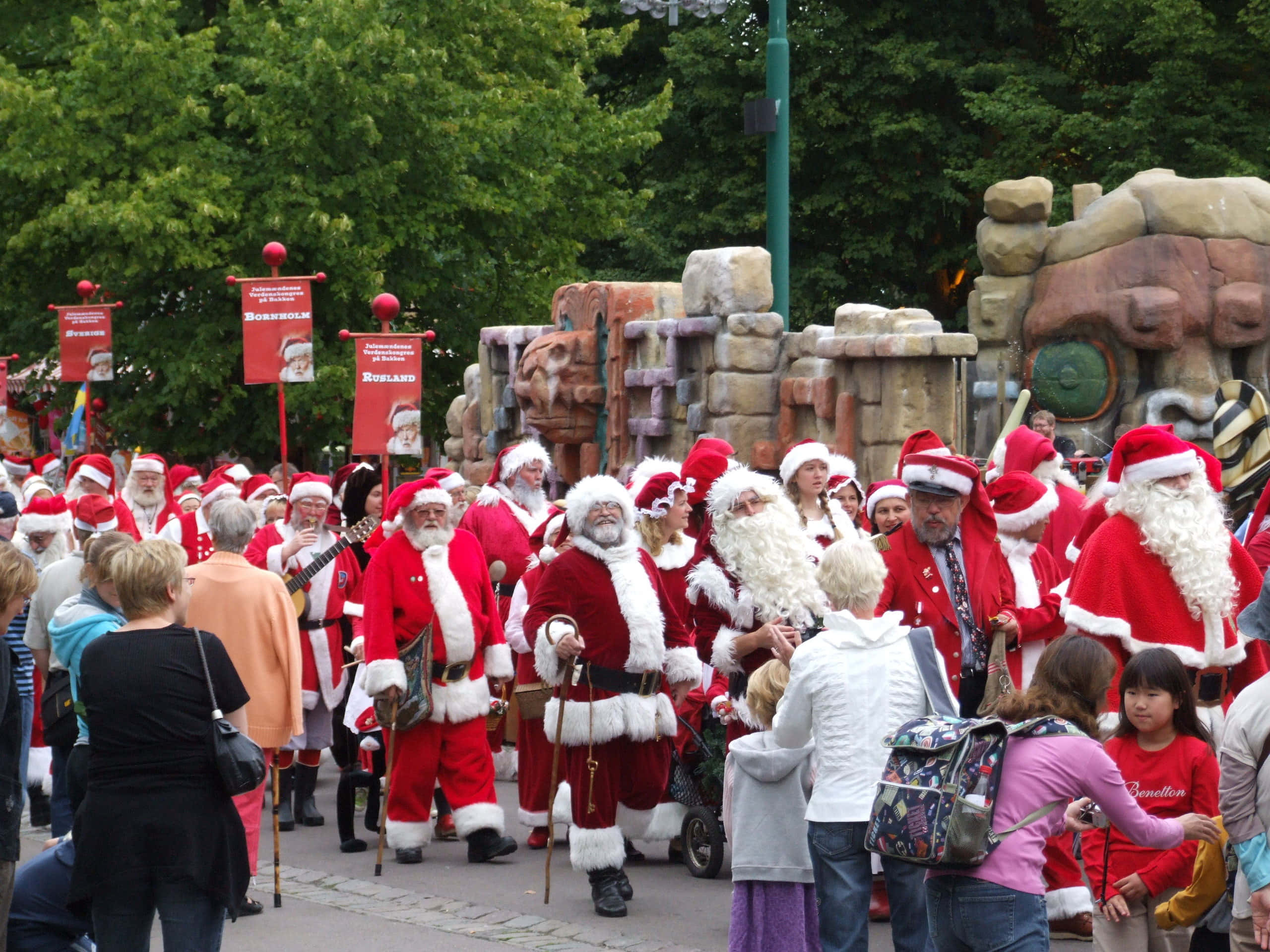 Santa Claus 桌布 壁紙 — 一場熱鬧的聖誕老人遊行，他們穿著紅色服裝，留著白鬍子，穿過一個有觀眾和主題石結構的節日戶外活動。