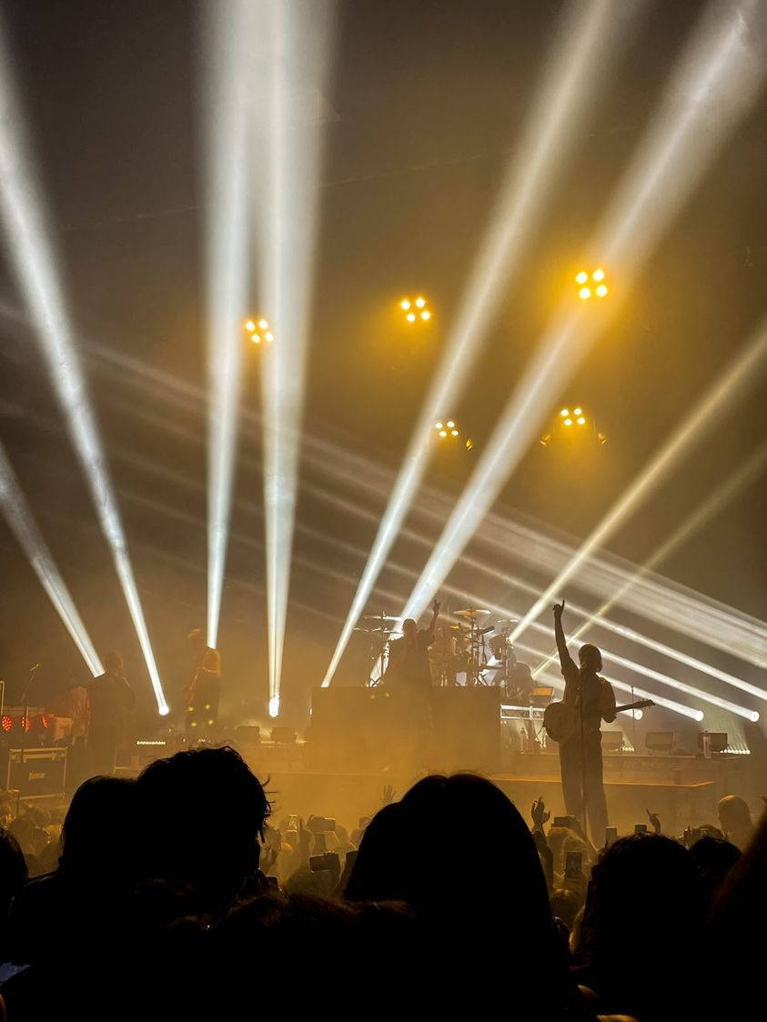 Chase Atlantic 桌布 壁紙 — 一場現場音樂會，舞台上的音樂家沐浴在穿過煙霧的白黃色戲劇性光束中，被熱情的人群剪影。