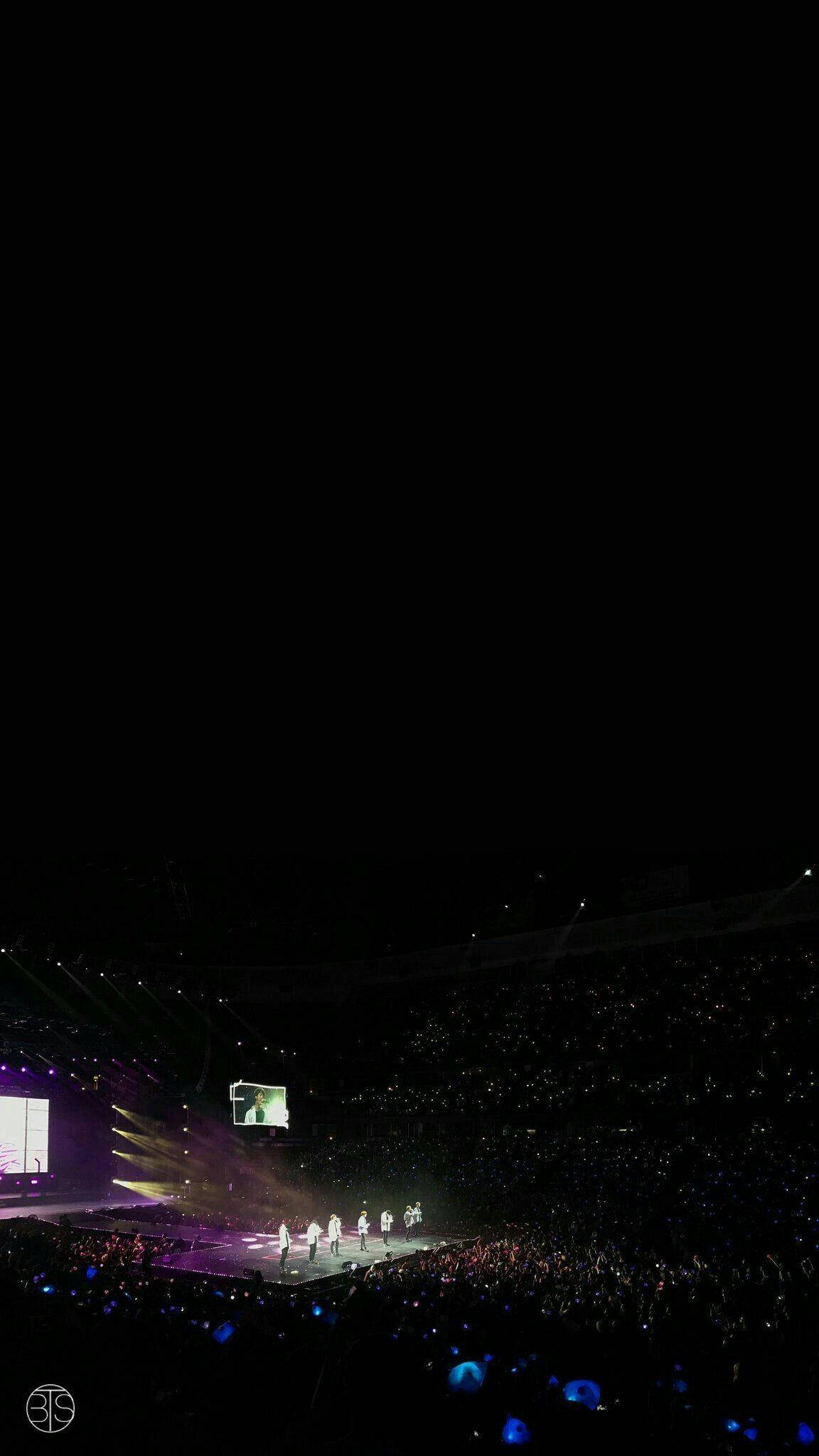 BTS 黑色 桌布 壁紙 — 一場現場音樂會，表演者在明亮照明的舞台上，在充滿觀眾發出藍色手機燈光的黑暗場館中演出。