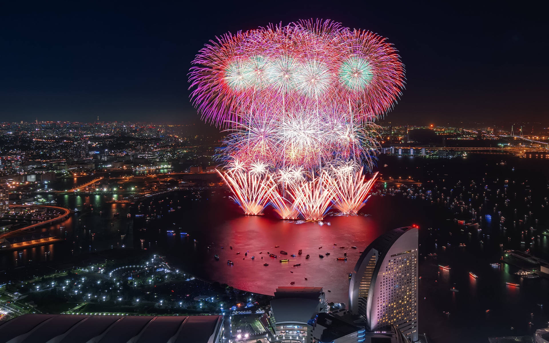 Yokohama 桌布 壁紙 — 一場絢麗的煙火表演在夜晚閃爍的城市海濱上空綻放，以鮮豔的紅色、紫色和綠色照亮黑暗的天空。